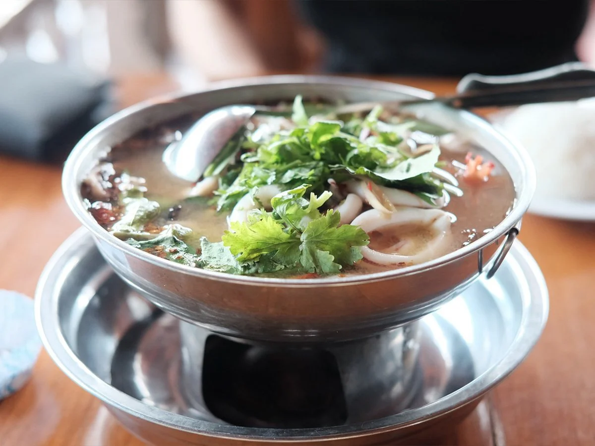 Bowl of Asian noodle soup with greens, herbs, and seafood, served in a metal bowl on a wooden table.