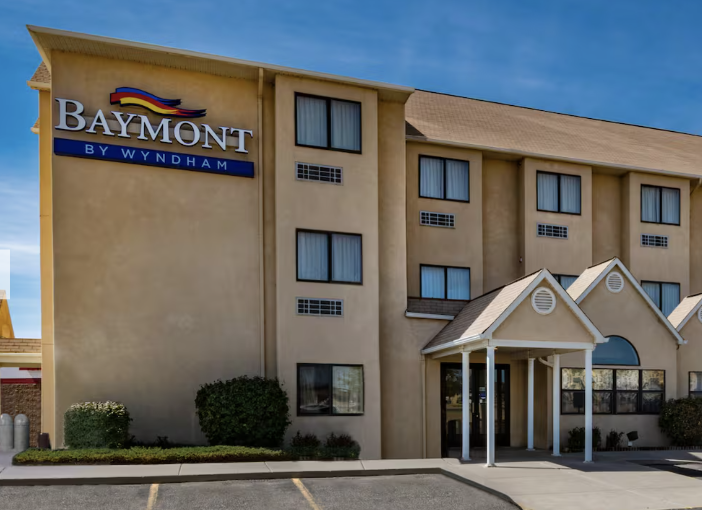 Exterior of Baymont by Wyndham hotel with beige stucco walls, multiple windows, and a small entrance with white pillars, under a blue sky.