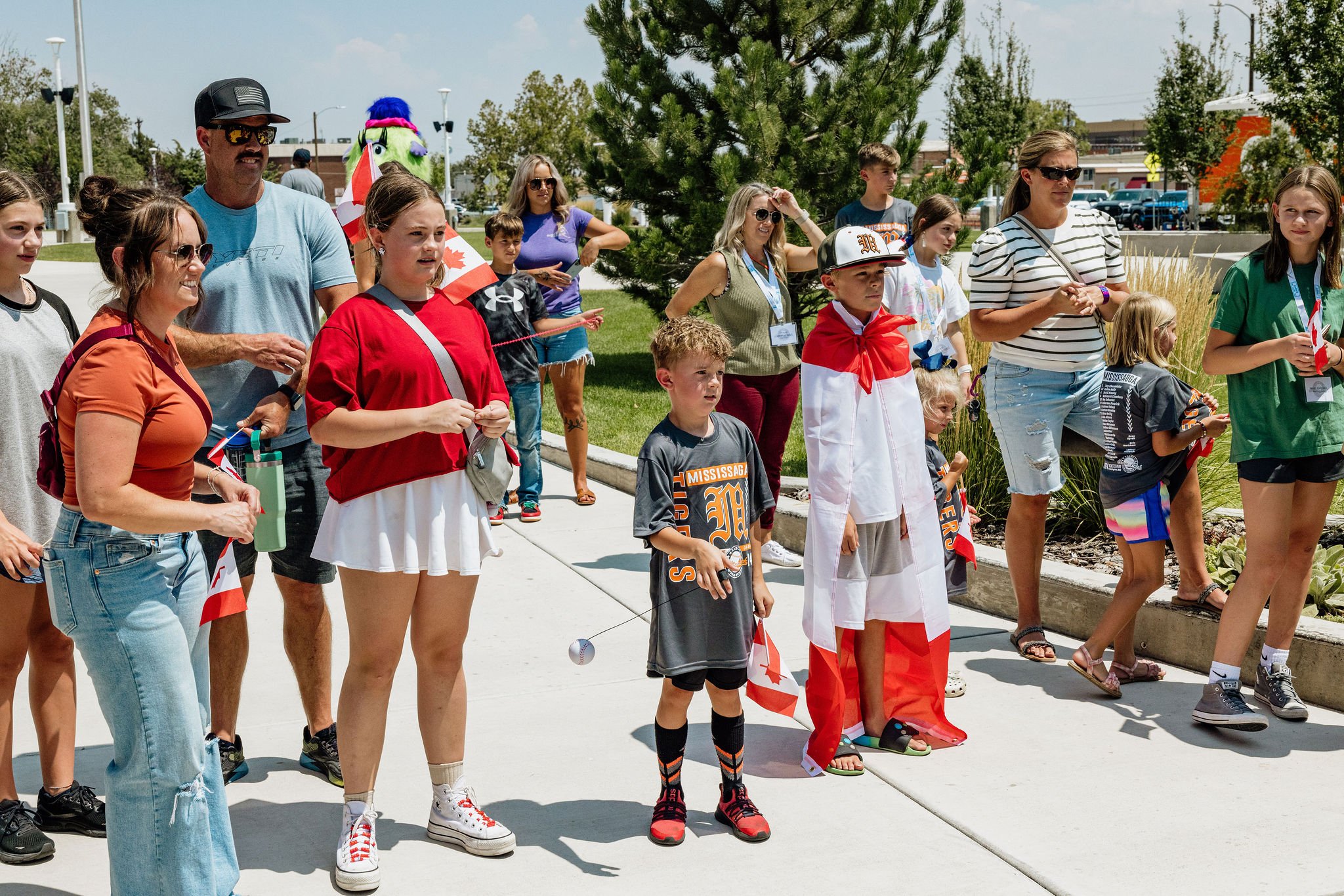 A group of people, including children and adults, gathered outdoors on a sunny day, some holding Canadian flags. They appear to be participating in or watching an event, with a colorful mascot in the background and trees and buildings nearby.