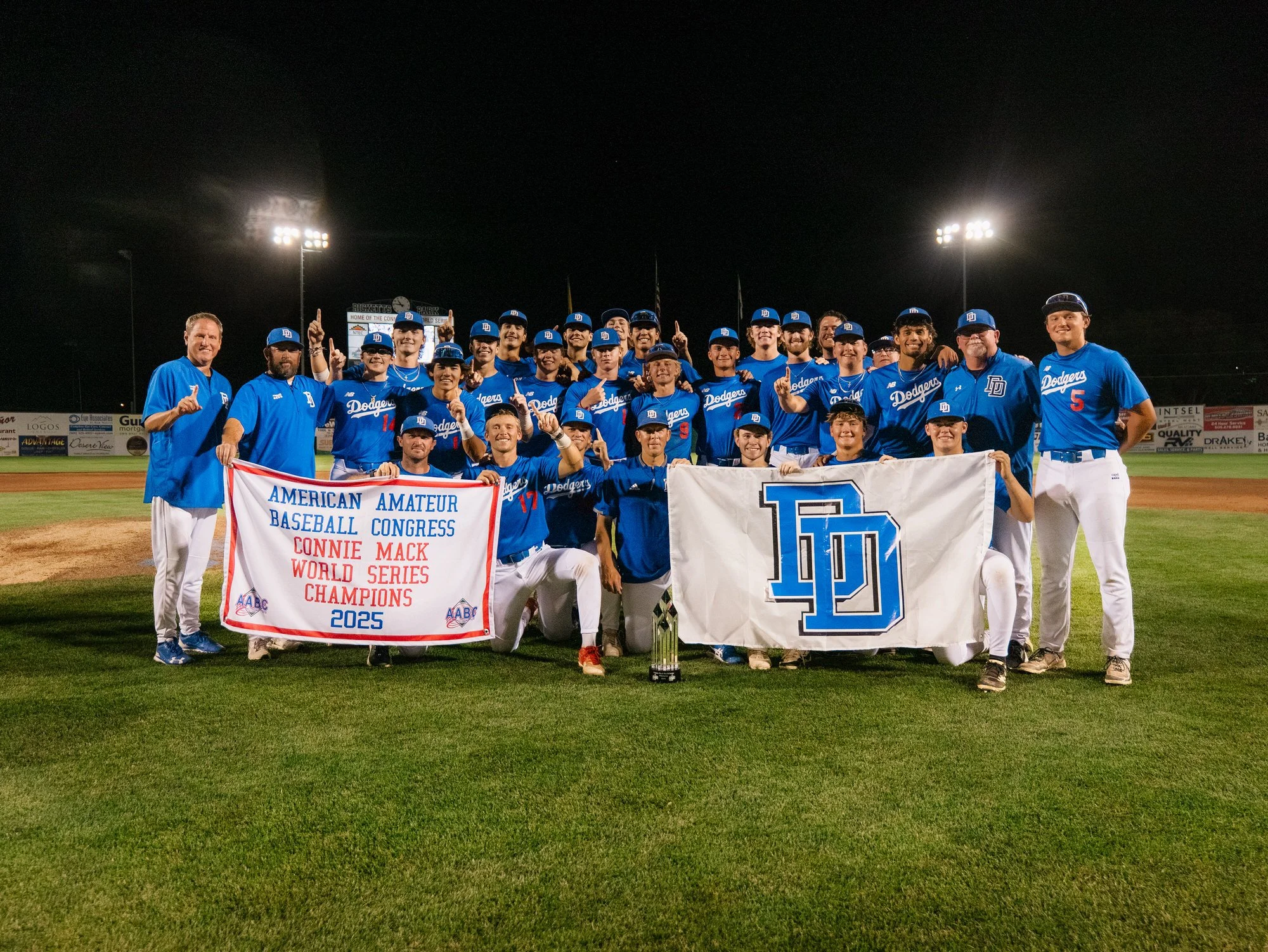 A group of baseball players and coaches on the field at night, celebrating their championship victory. They are wearing blue uniforms with 'Dodgers' written on them, and some are holding a banner that reads 'American Amateur Baseball Congress, Baseball Congress, Connie Mack, World Series Champions 2025.' They are smiling, with some raising one finger in the air, and a trophy is placed on the ground in front of them.
