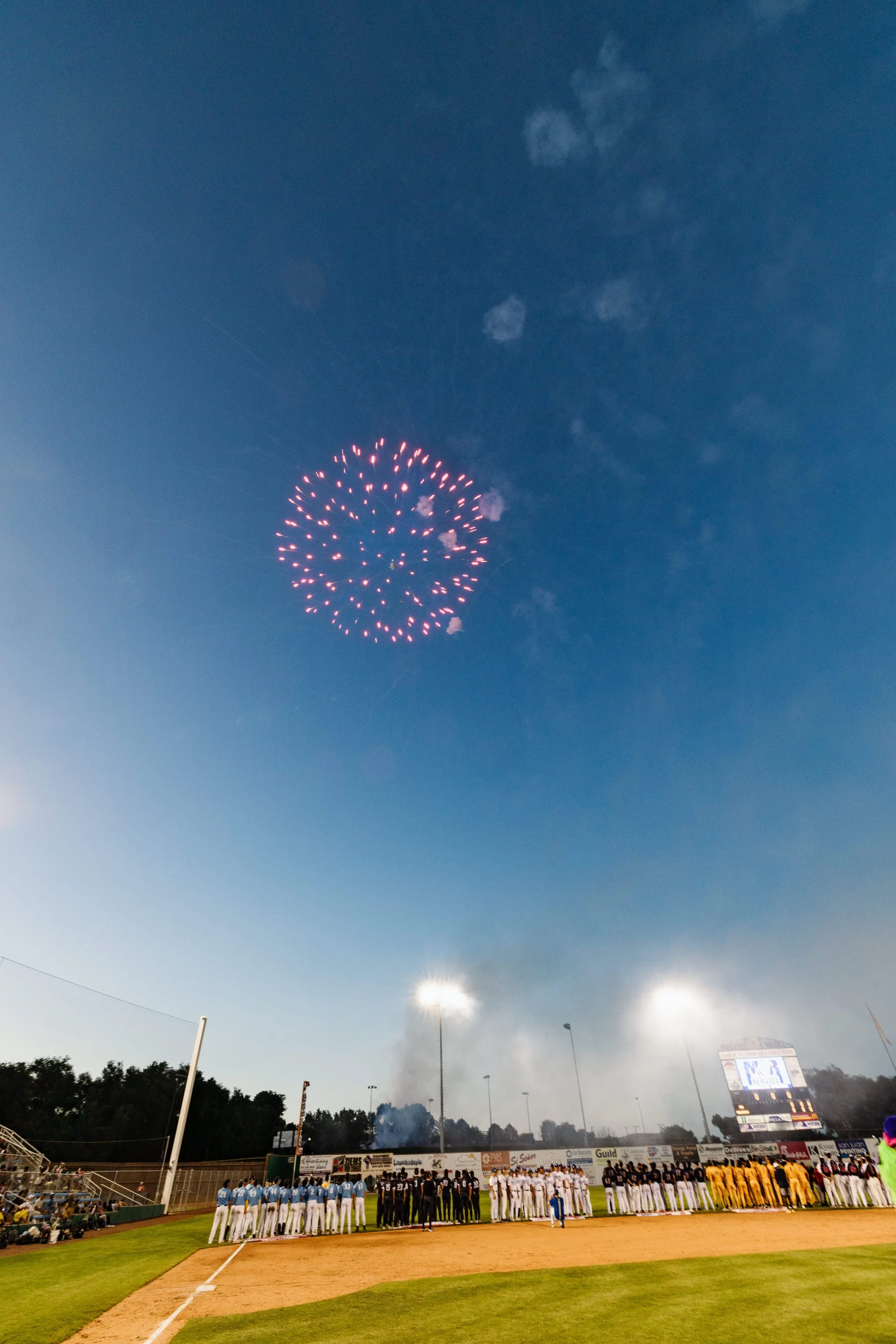 Baseball players standing in two lines on the field, facing each other, with fireworks in the evening sky above stadium. Spectators and scoreboard visible in the background.