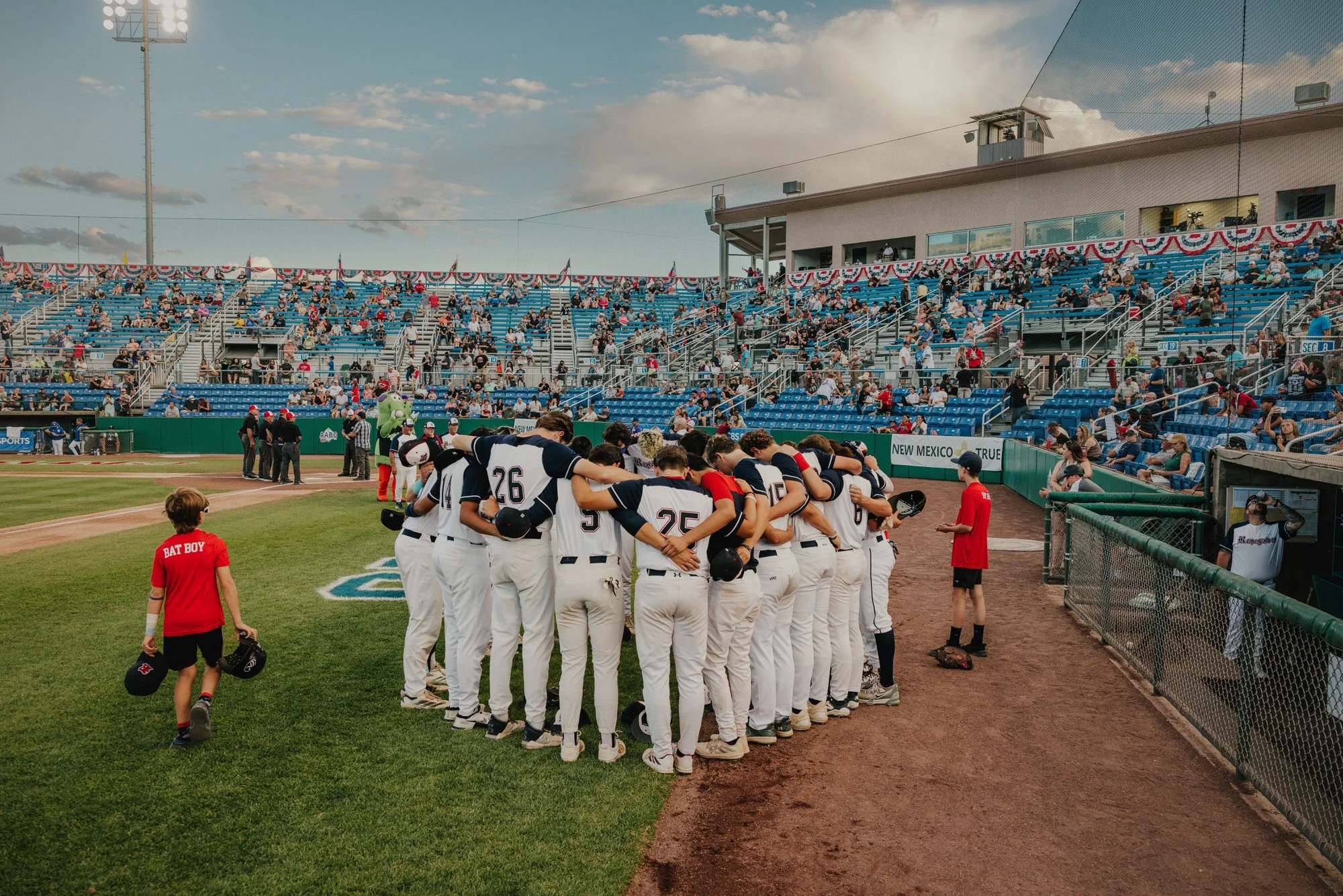 A baseball team dressed in white uniforms huddles together on the field, likely during a game or practice. They are standing close with their arms around each other's shoulders. Several young children walk nearby, one wearing a red shirt labeled 'Bat Boy'. The stadium seats are filled with spectators, and there are bright lights and a clear sky overhead.