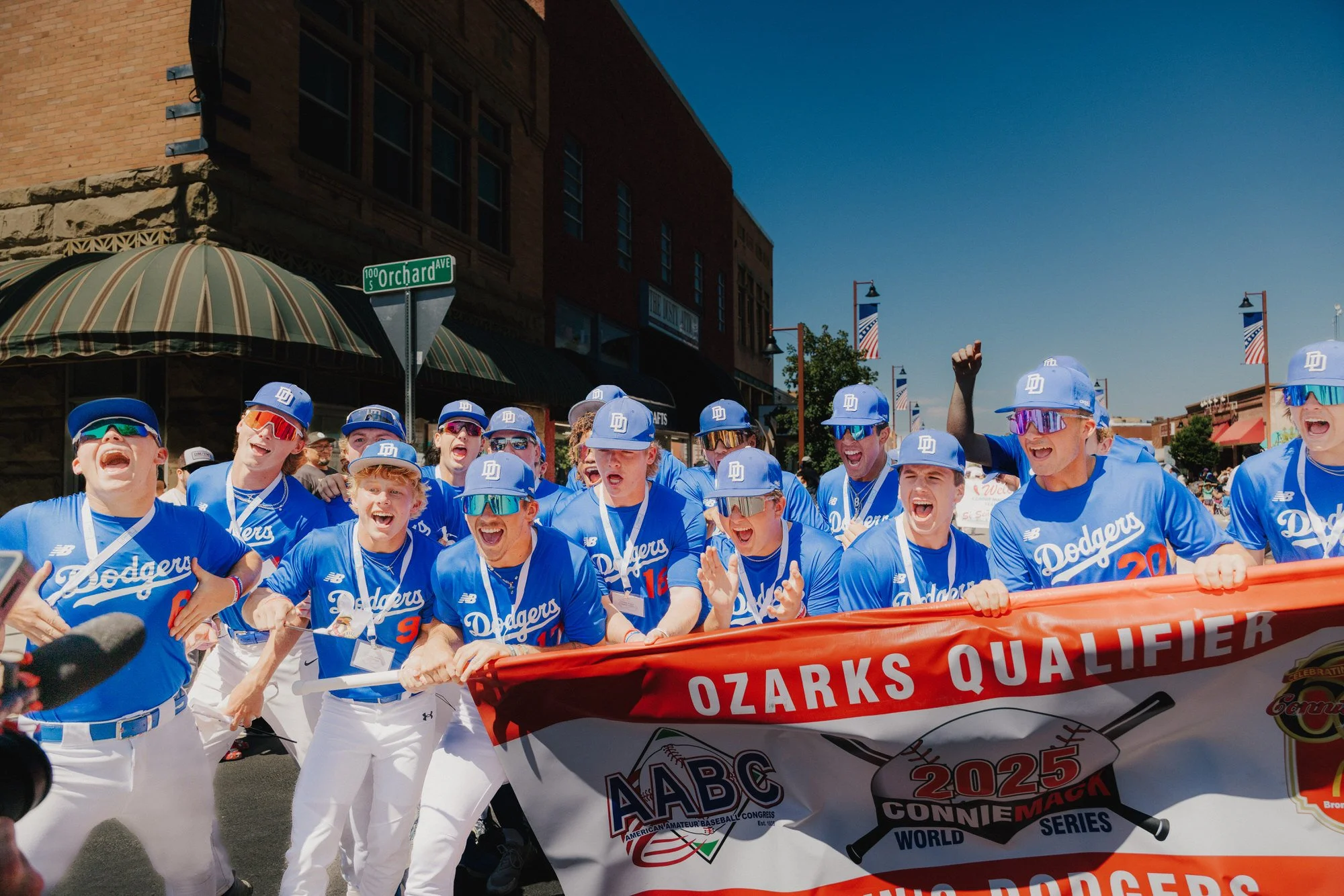 A group of cheering baseball players in blue uniforms and white pants participating in a parade, holding a banner that reads 'Ozarks Qualifier' and celebrating their team achievements.