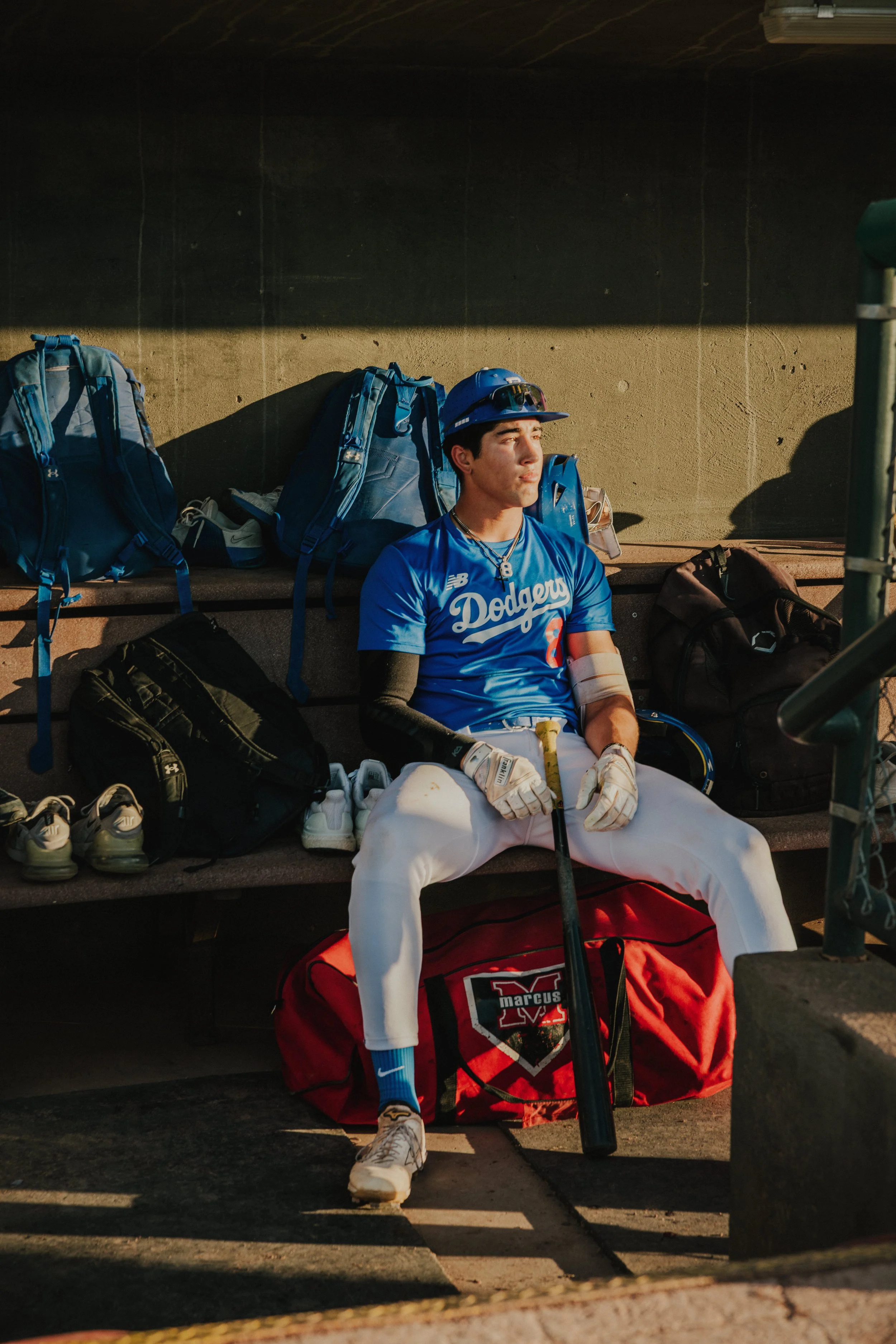 A young baseball player in uniform sitting on a bench, wearing a blue Dodgers jersey, white pants, and a blue helmet, surrounded by sports bags and equipment.