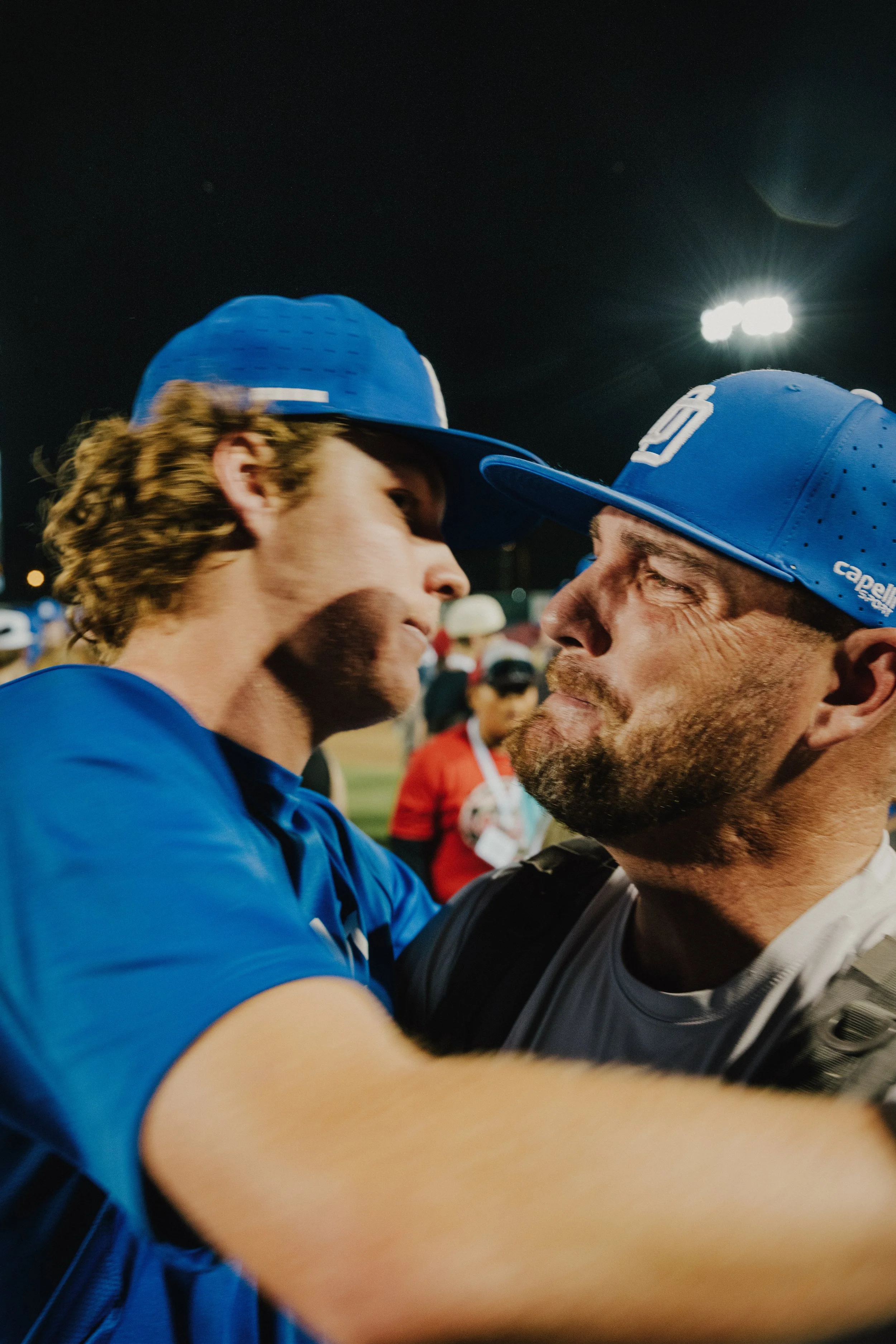 Two men wearing blue caps and shirts, face to face, close up, at a sporting event at night, with bright stadium lights in the background.