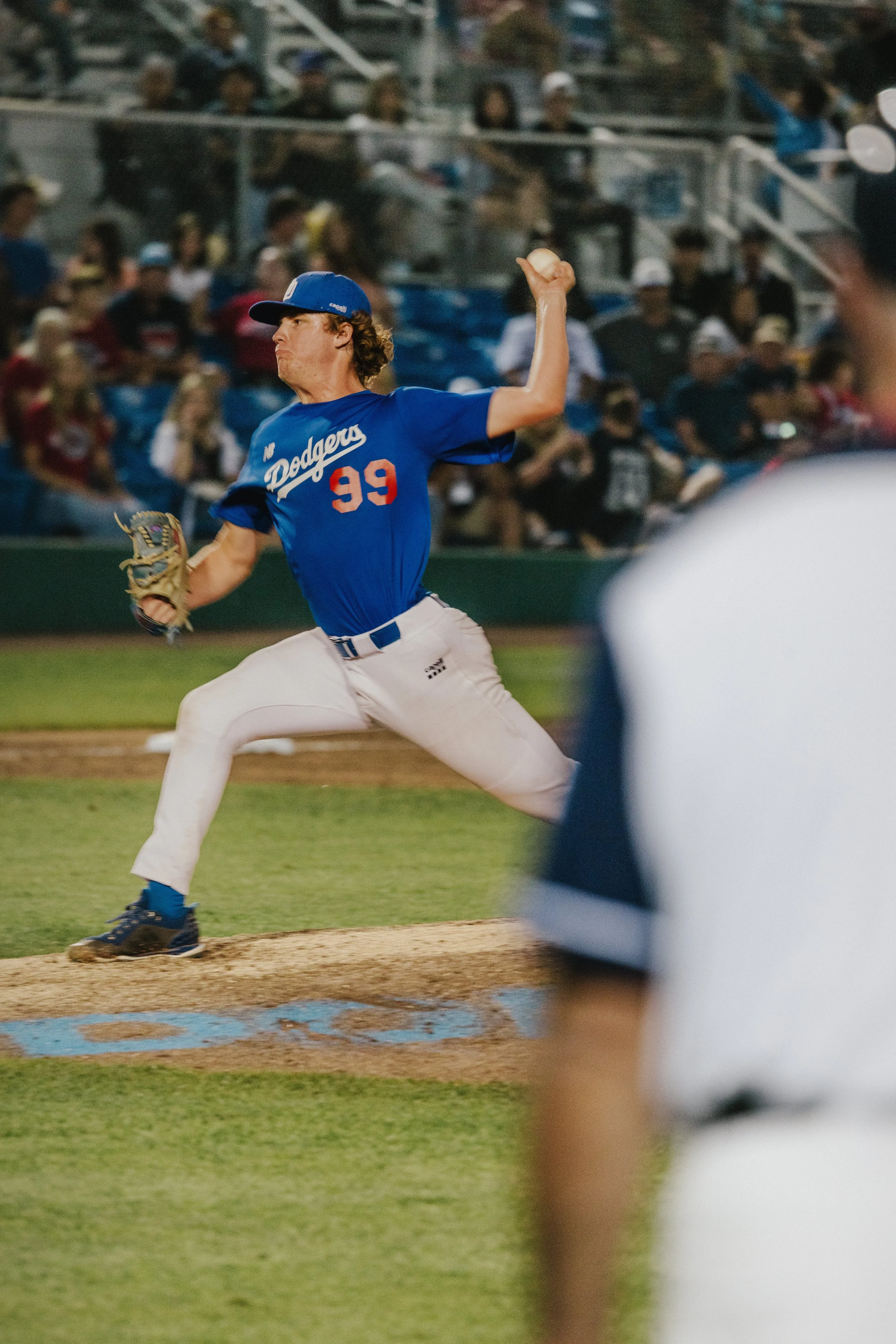A baseball pitcher in a blue uniform and blue cap with the name "Dodgers" and the number 99 throws a pitch on a baseball field during a game with spectators in the stands.