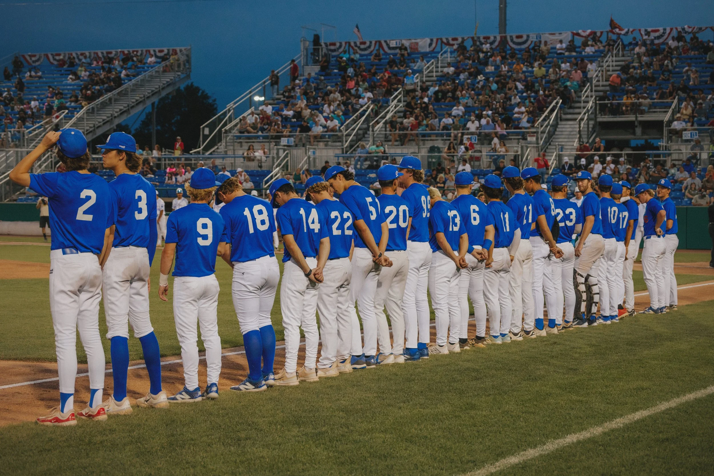 Baseball team in blue jerseys and white pants standing on the field, with some players adjusting hats, while a crowd sits in stadium seats in the background during the evening.