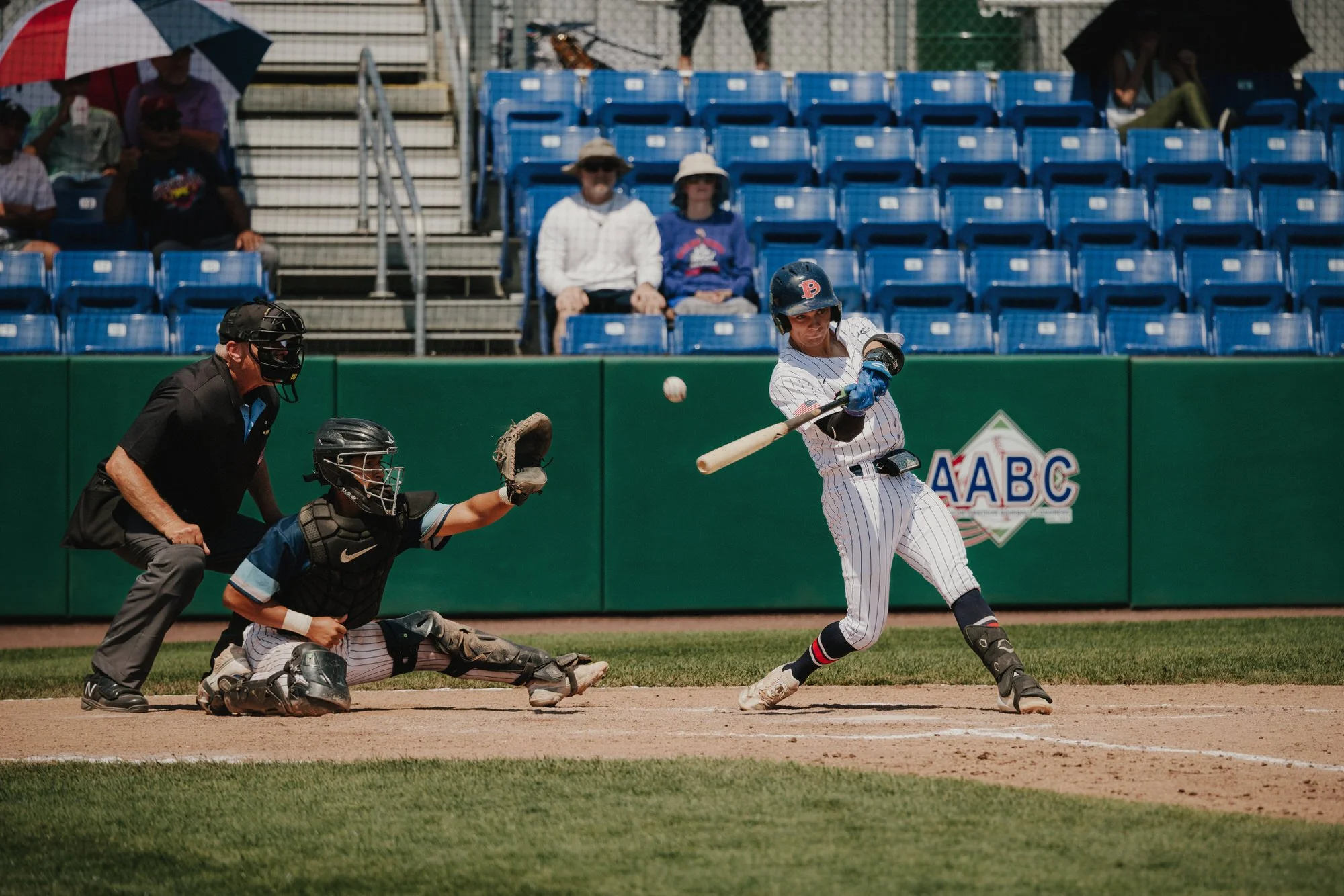 A baseball player batting as the catcher and umpire prepare for the play, with spectators seated in the background at the stadium.