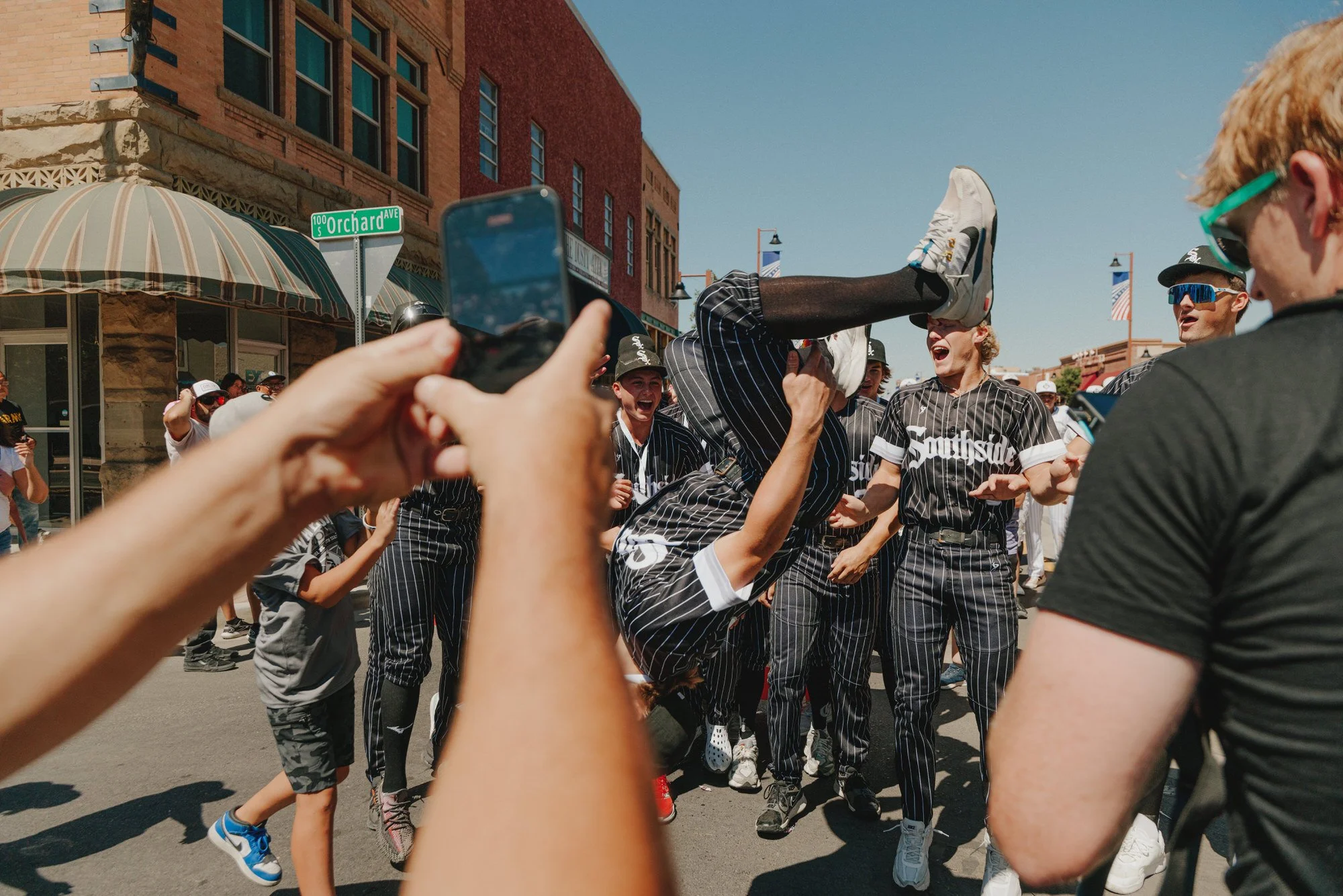 A street scene with people celebrating, some wearing baseball jerseys. One person is holding another upside down, and there's a person taking a photo with their phone. The background shows storefronts, a street sign for Orchard Avenue, and banners hanging on street lamps.