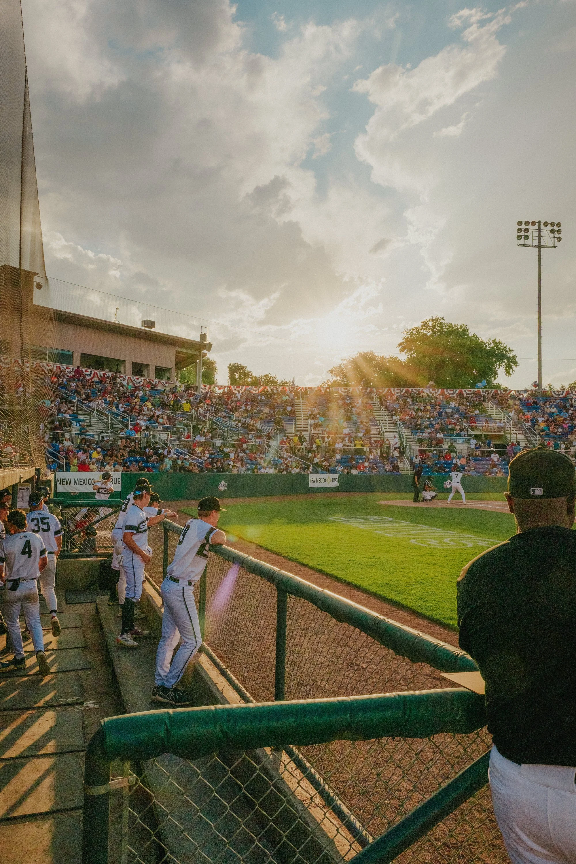 Baseball field during game with players, spectators in stands, and cloudy sky with the sun shining.