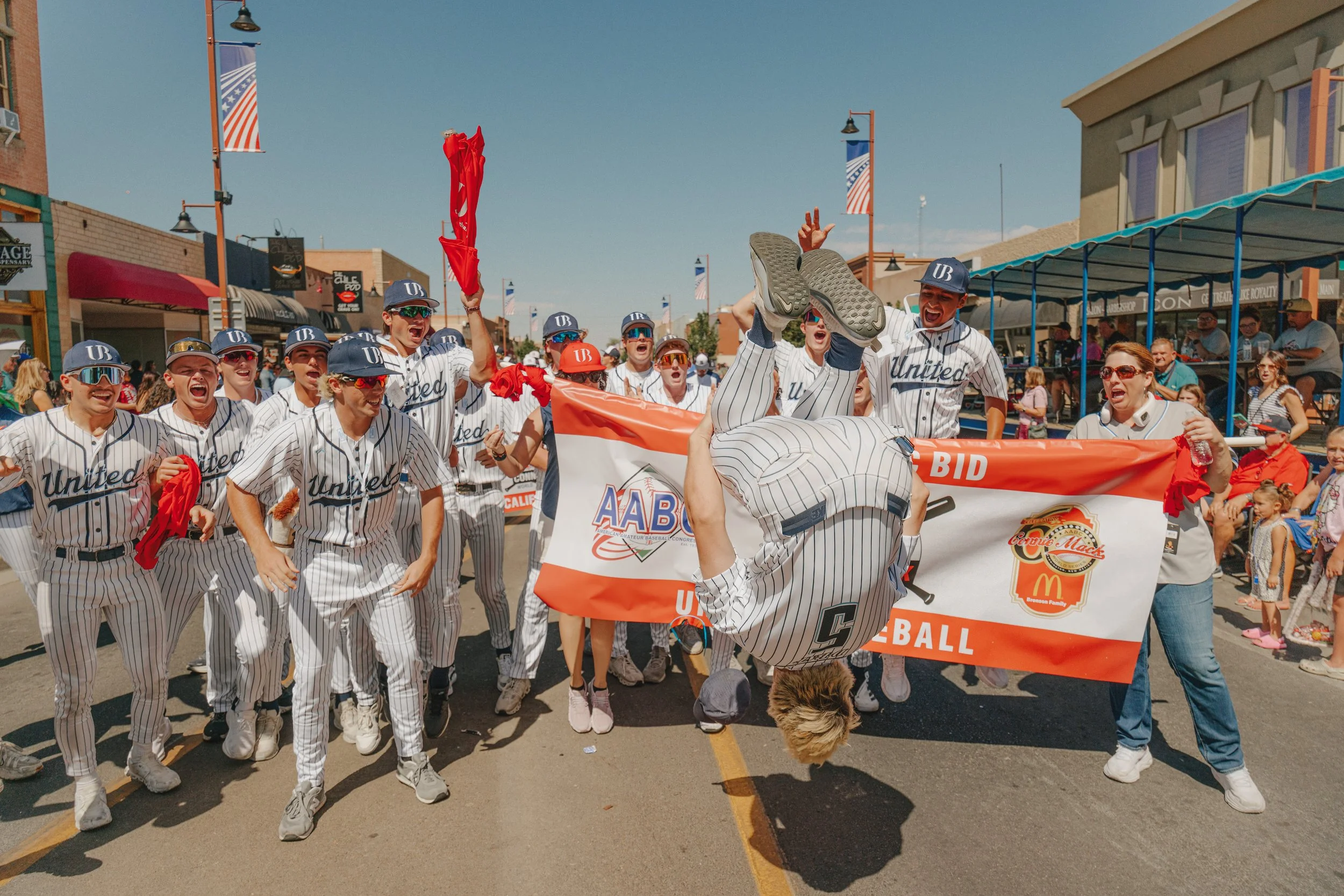 Group of young baseball players wearing jerseys with 'United' and caps, celebrating and cheering during parade on a street with shops and American flags.