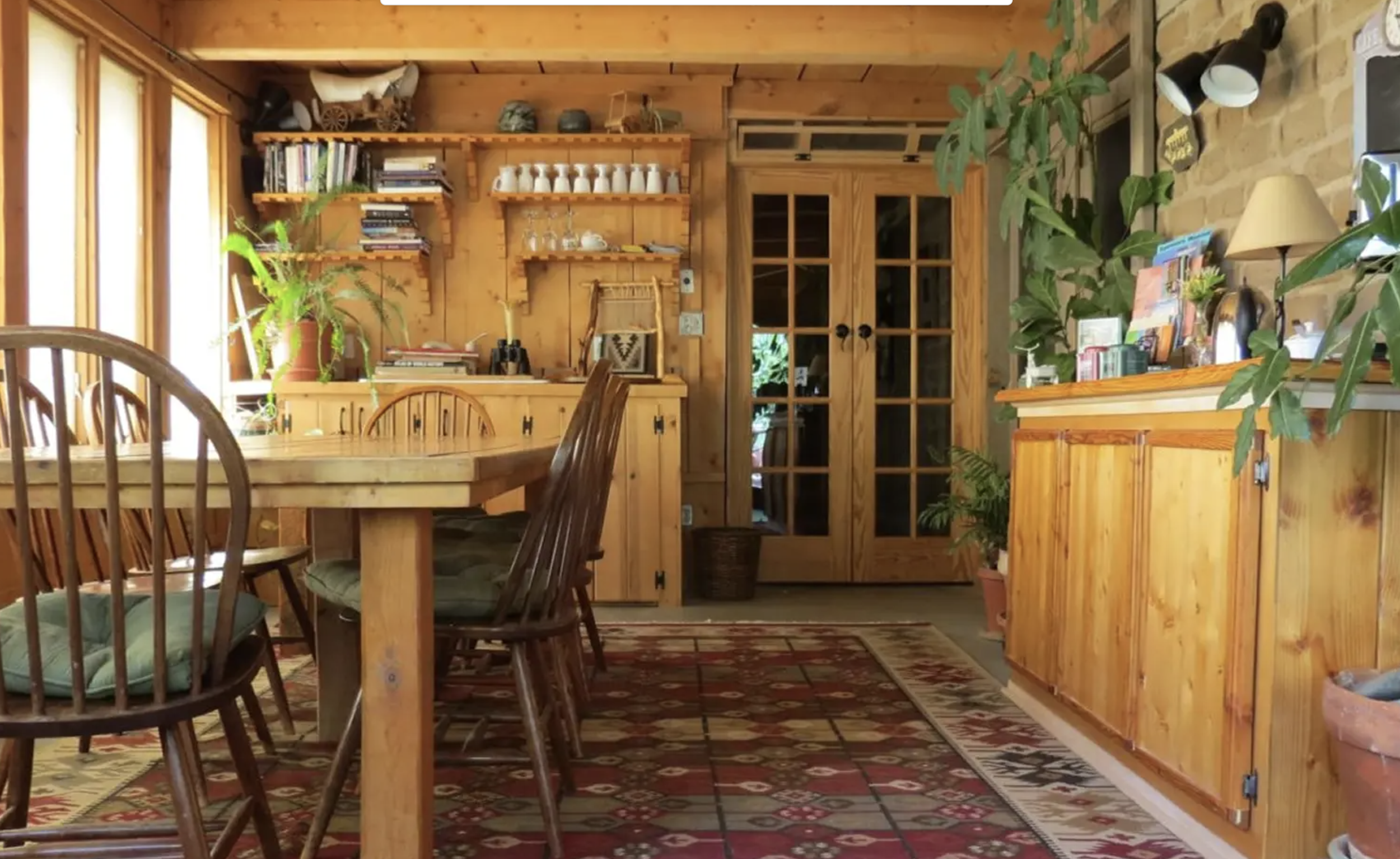 A cozy kitchen with wood-paneled walls, a wooden dining table with chairs, shelves with books and dishes, and various plants. There are double glass-paneled wooden doors and a patterned rug on the floor.