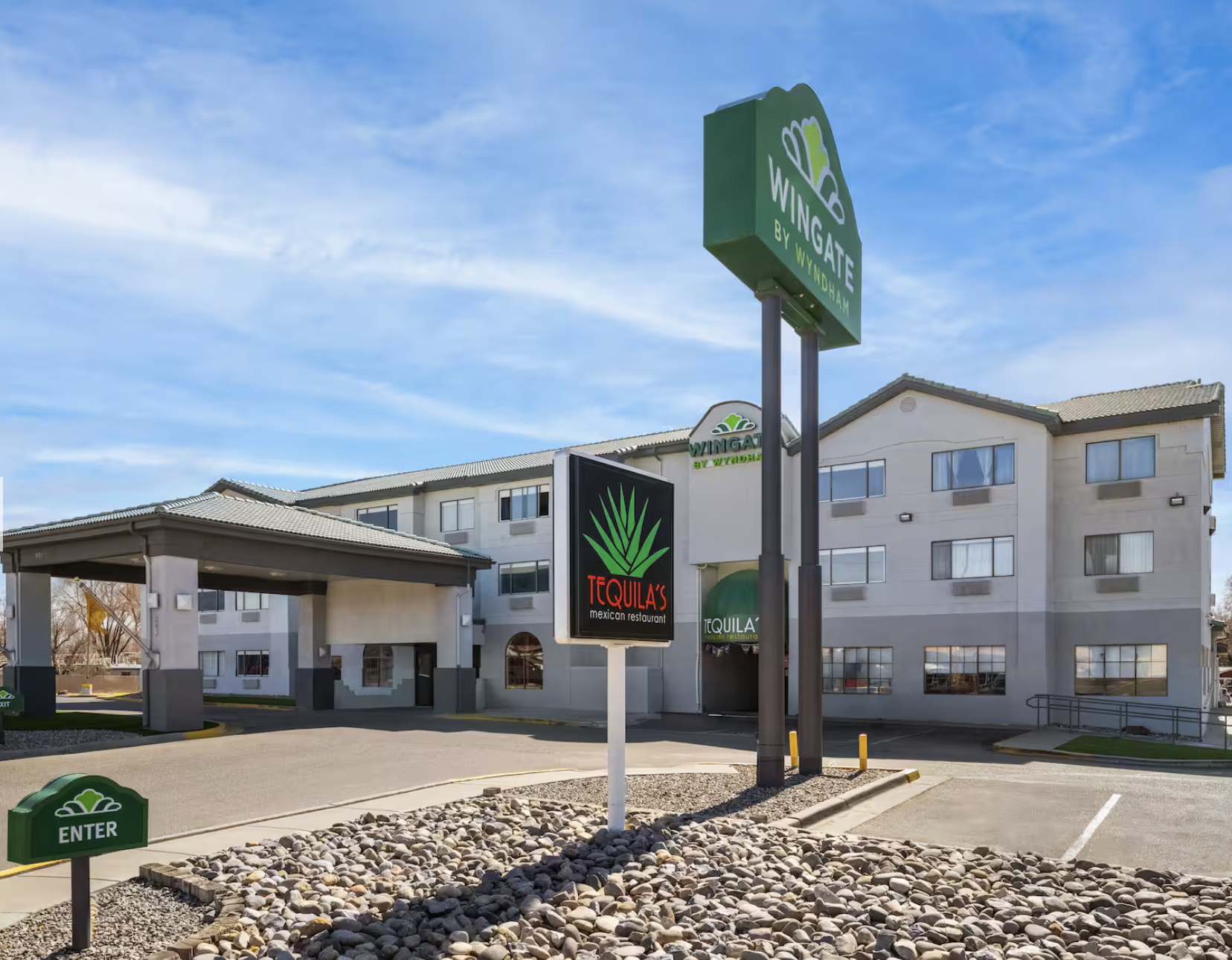 Exterior view of a hotel with signs for WINGATE by WYNDHAM and a Mexican restaurant called TEQUILA'S. There is a covered entrance, parking lot, and a landscaped area with rocks and an 'ENTER' sign. The building is four stories tall with numerous windows, under a blue sky with some clouds.
