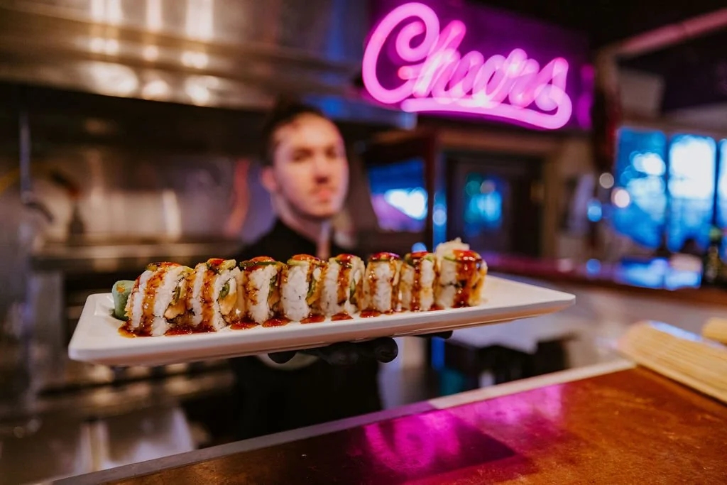 A server presenting a plate of sushi rolls with sauce in a restaurant with neon signs in the background.