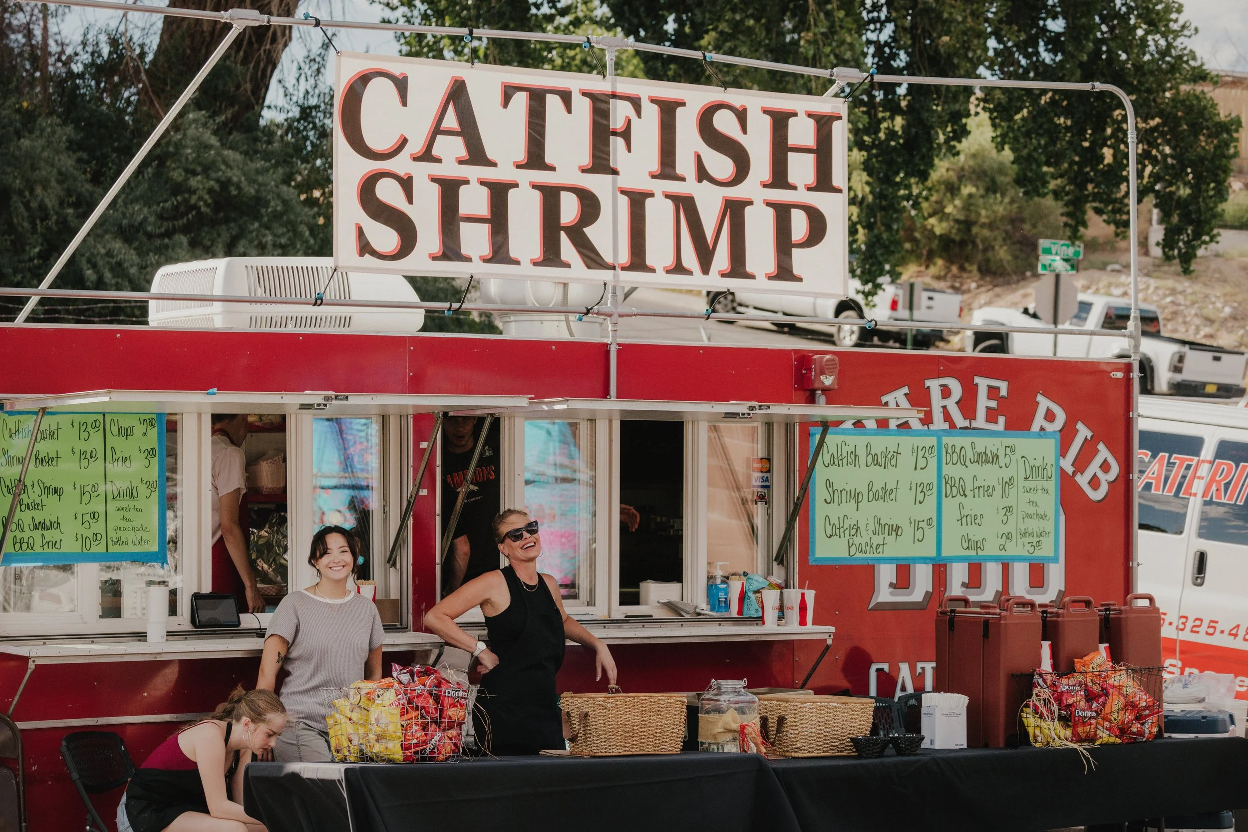A red food stand with a sign that reads 'CATCH FISH SHRIMP'. There are several people, including a woman with sunglasses and a young girl, smiling and working at the stand, with green menus listing seafood options like catfish baskets and shrimp baskets, and some trees and parked vehicles in the background.