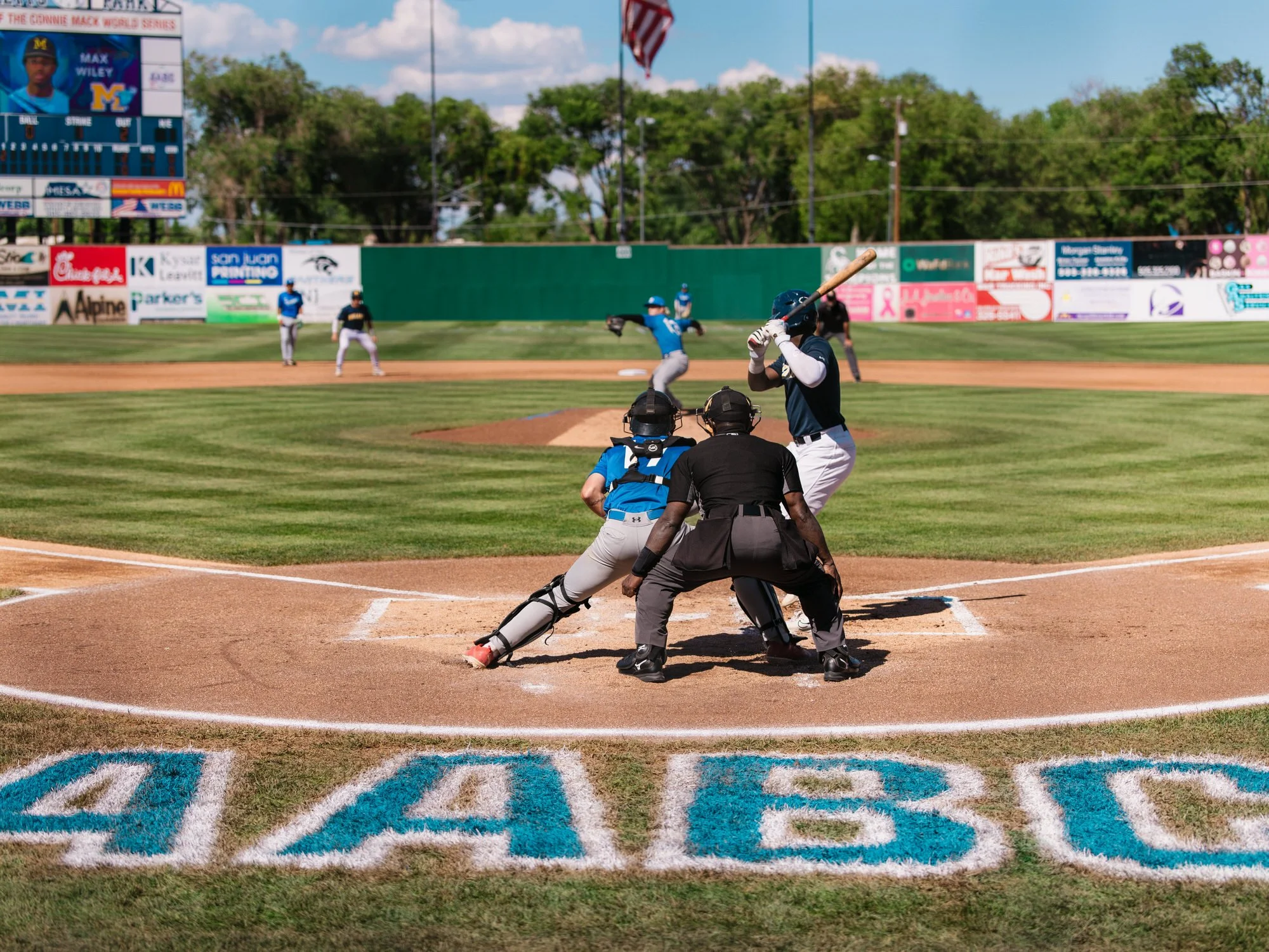 Baseball game at a stadium with players, officials, and a batter preparing to hit. The scoreboard and advertisements are visible in the background.