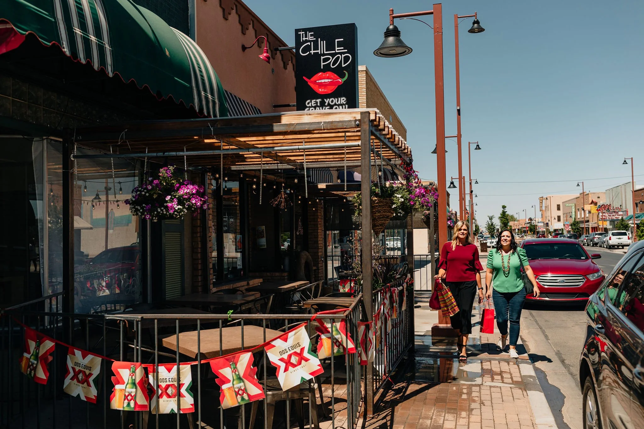 Street view with pedestrians, storefronts, banners, and hanging flower baskets on a sunny day.