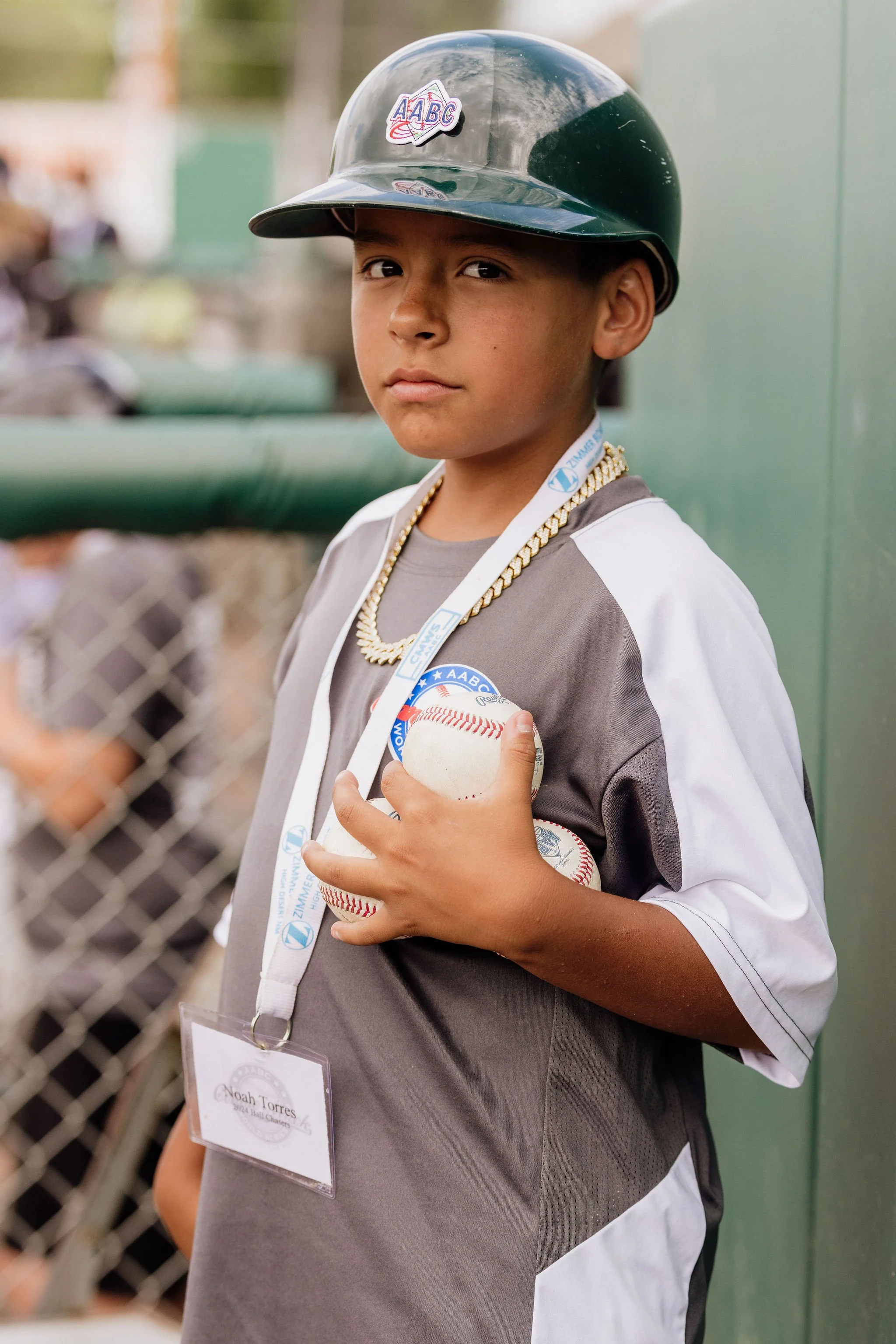 A young baseball player wearing a black helmet with an 'AABC' patch, holding a baseball against a chain-link fence.