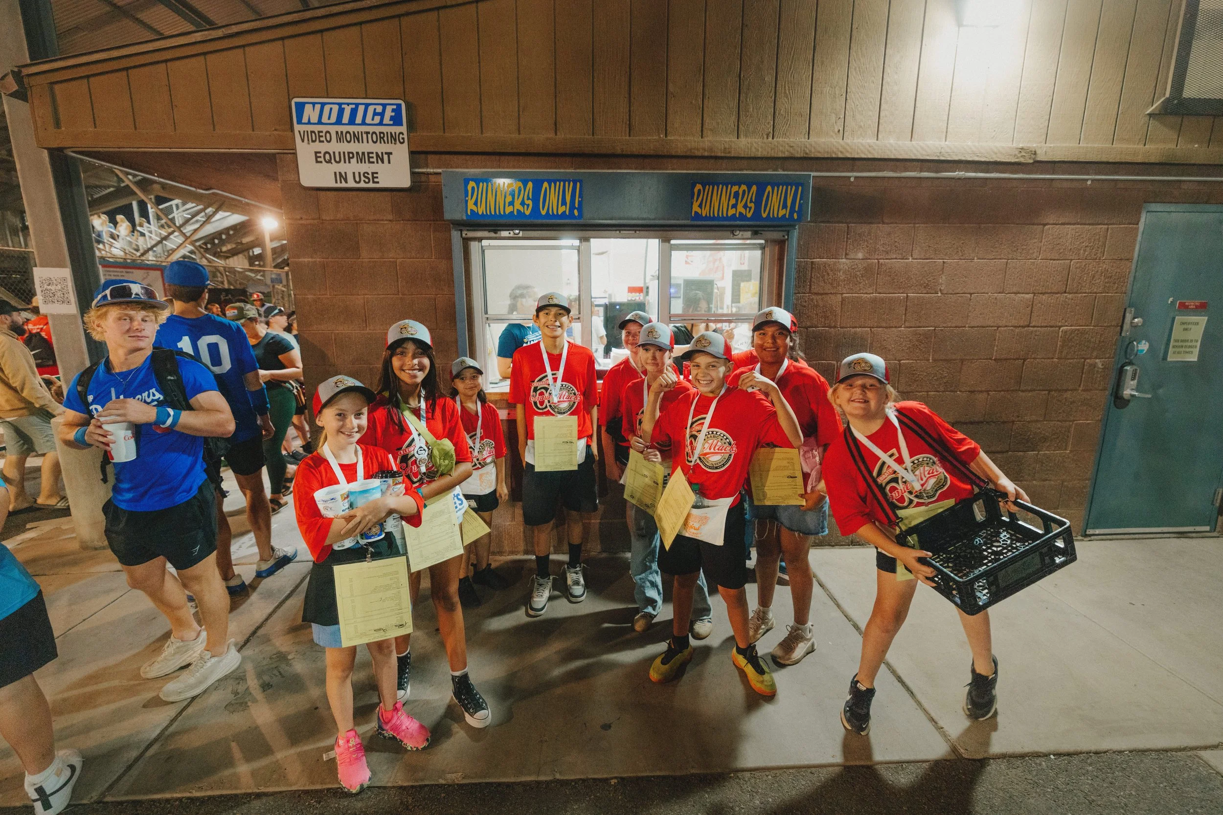 A group of young athletes, mostly girls, wearing red sports jerseys and gray caps with a logo, standing outside a building near a snack window labeled "Runners Only!" at a sports stadium. They are smiling, holding medals and papers, with some holding water bottles. There are people and stadium seating visible in the background.