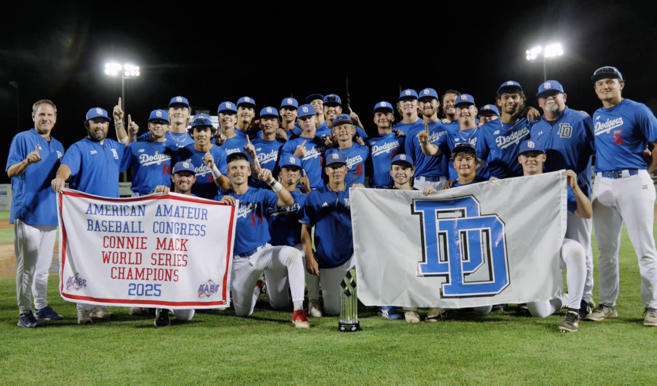 A group of baseball players and coaches on a field at night, celebrating their victory with banners that read 'American Amateur Baseball Congress, Connie Mack World Series Champions 2023' and a large trophy in front.