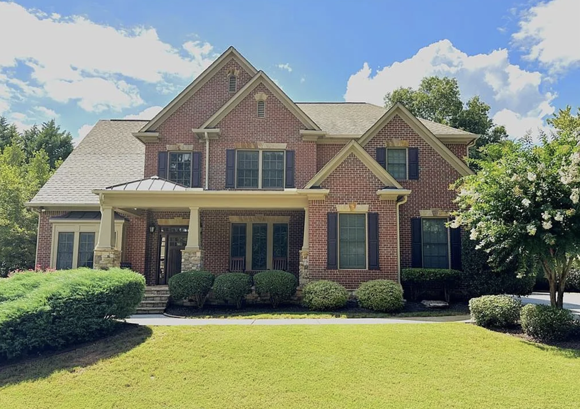 A large, two-story brick house with a front porch supported by columns and a gabled roof, surrounded by neatly trimmed bushes and trees, under a partly cloudy sky.