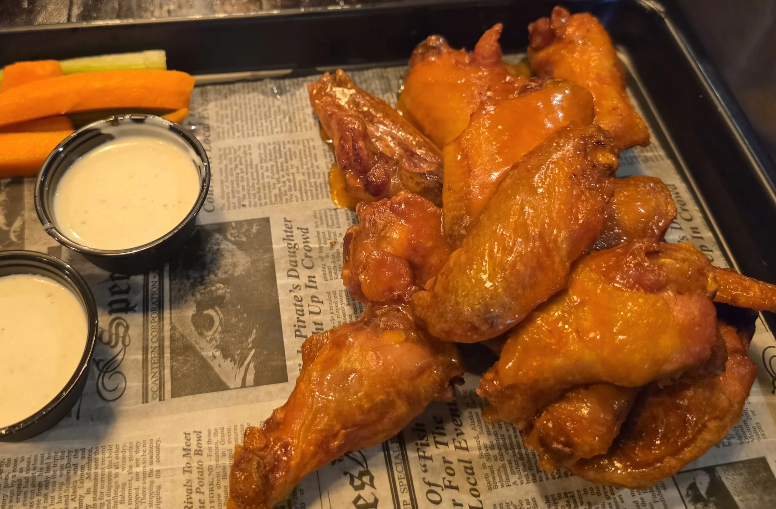 Buffalo chicken wings with orange sauce served on newspaper, with side of carrot and celery sticks and two small cups of dipping sauce.