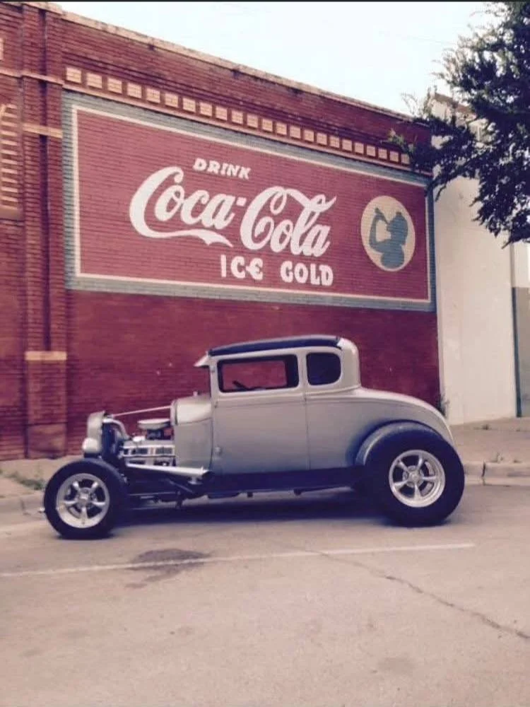 Vintage car parked in front of a brick building with a Coca-Cola advertisement on the wall.