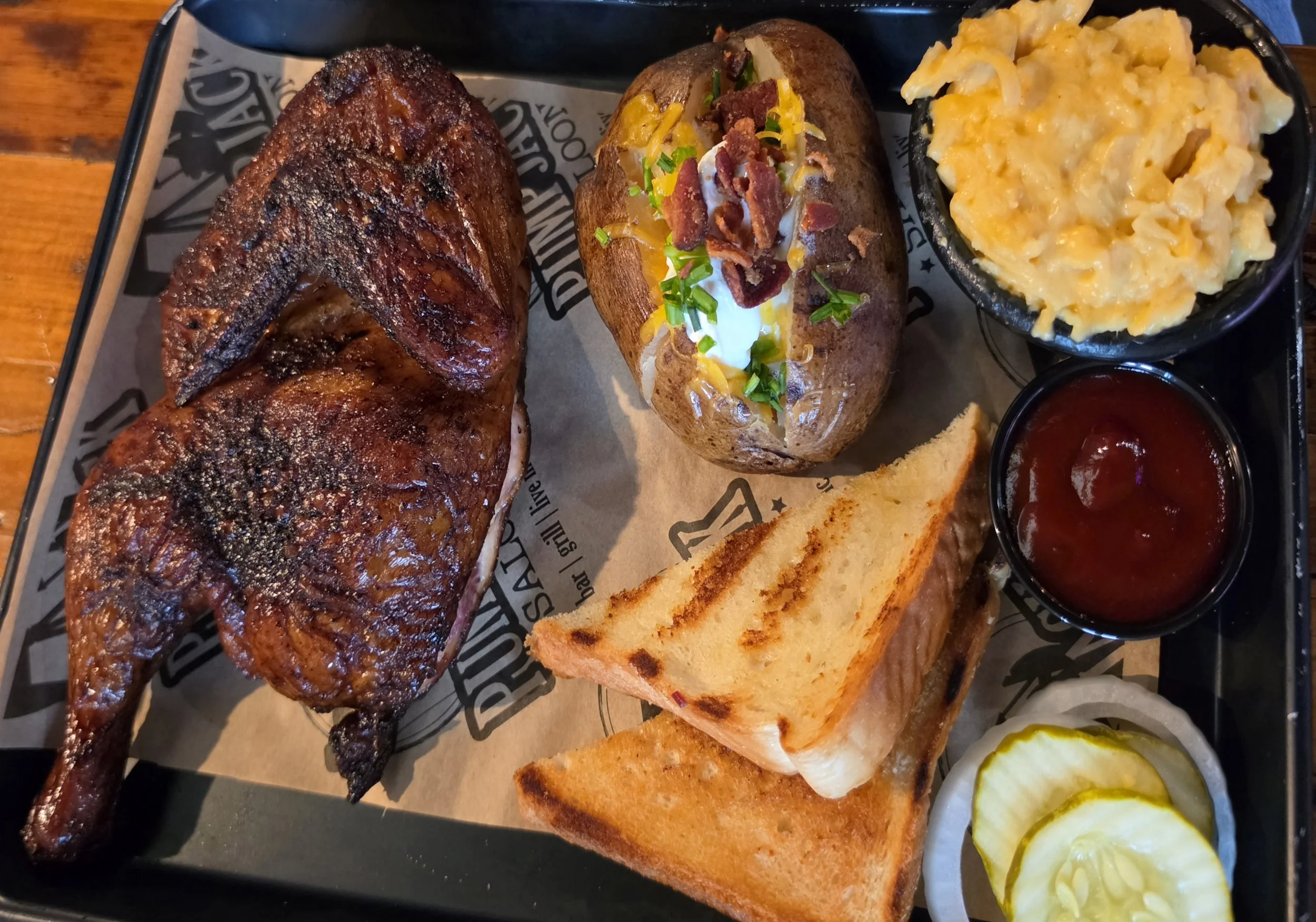 A serving tray with a roasted chicken leg, a loaded baked potato with cheese, sour cream, and bacon, a slice of garlic bread, and pickles with mustard.