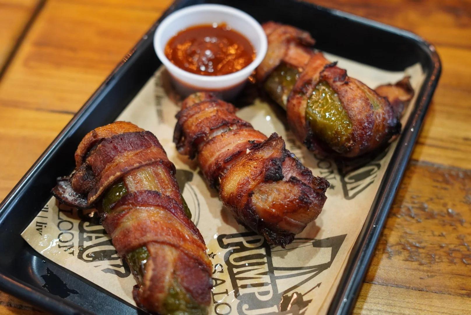 Bacon-wrapped jalapeño poppers with a side of spicy dipping sauce on a black serving tray.