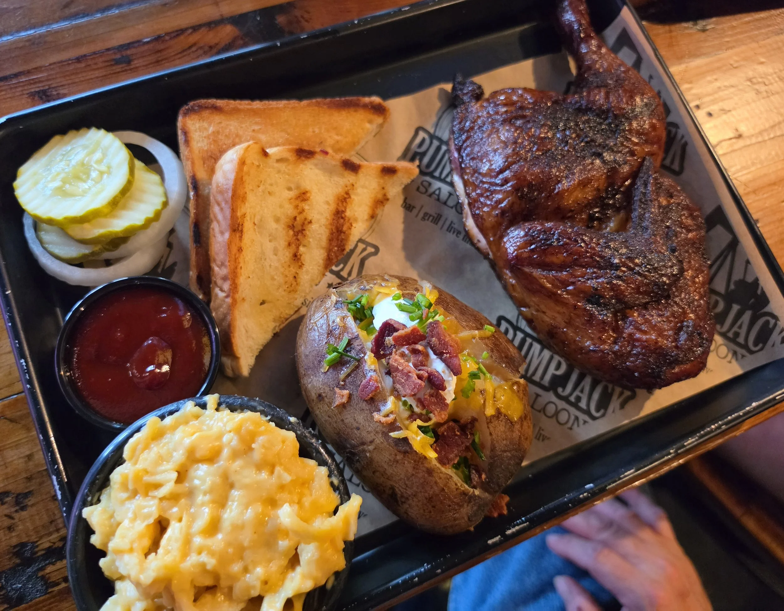 A black tray with a serving of mashed potatoes, toast, pickles, barbecue chicken, baked potato with sour cream and bacon bits, and a small container of ketchup.