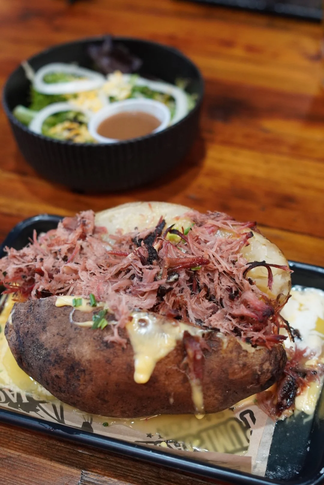 Baked potato topped with shredded pork, melted cheese, and chopped chives, served on a black tray. In the background, there is a salad with lettuce, onions, shredded cheese, and dressing in a black bowl, on a wooden table.