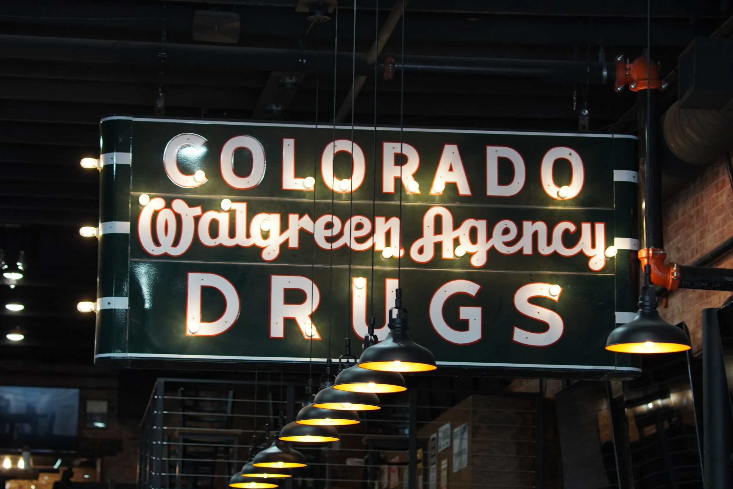 A sign with the words 'Colorado', '@talegreen.agency', and 'Drugs', with illuminated bulbs hanging below.