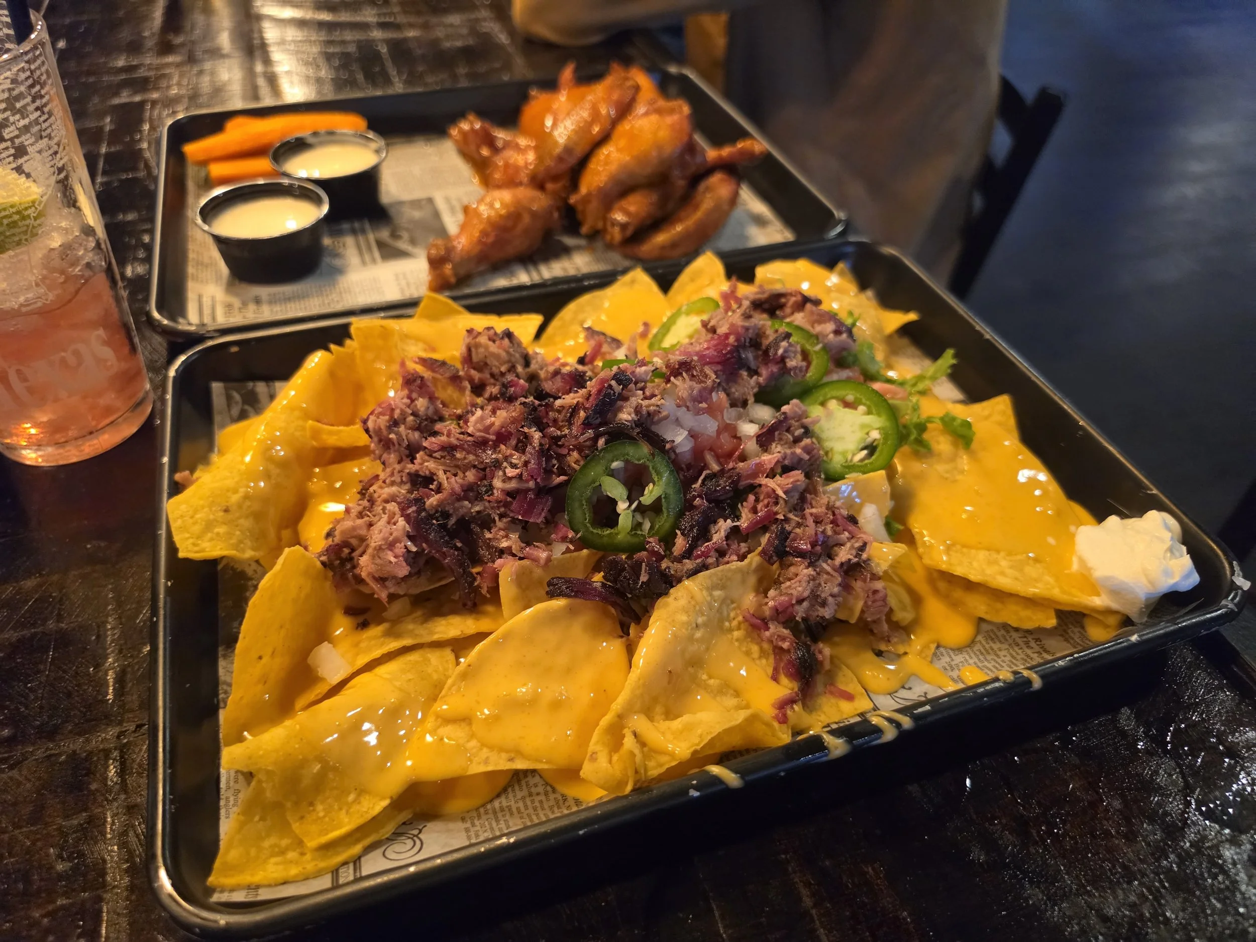 A tray of loaded nachos topped with shredded beef, sliced jalapeños, diced onions, and shredded cheese, with tortilla chips around the toppings. In the background, a separate tray of fried chicken wings, carrot sticks, and dipping sauces. A glass of pink-colored beverage is partially visible on the left.