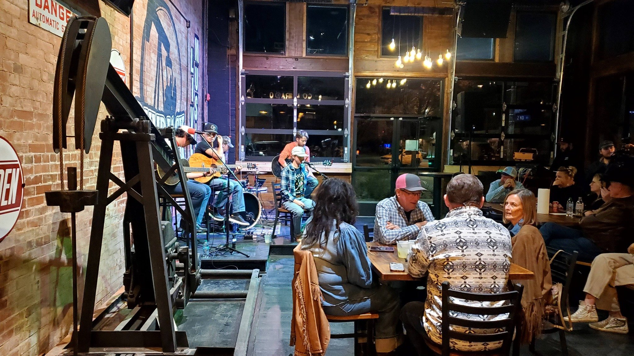 A live music performance at a restaurant or bar with patrons sitting at tables, some wearing hats, enjoying the evening. The band on stage has three musicians playing guitars, with musical equipment set up around them. The interior features brick wal