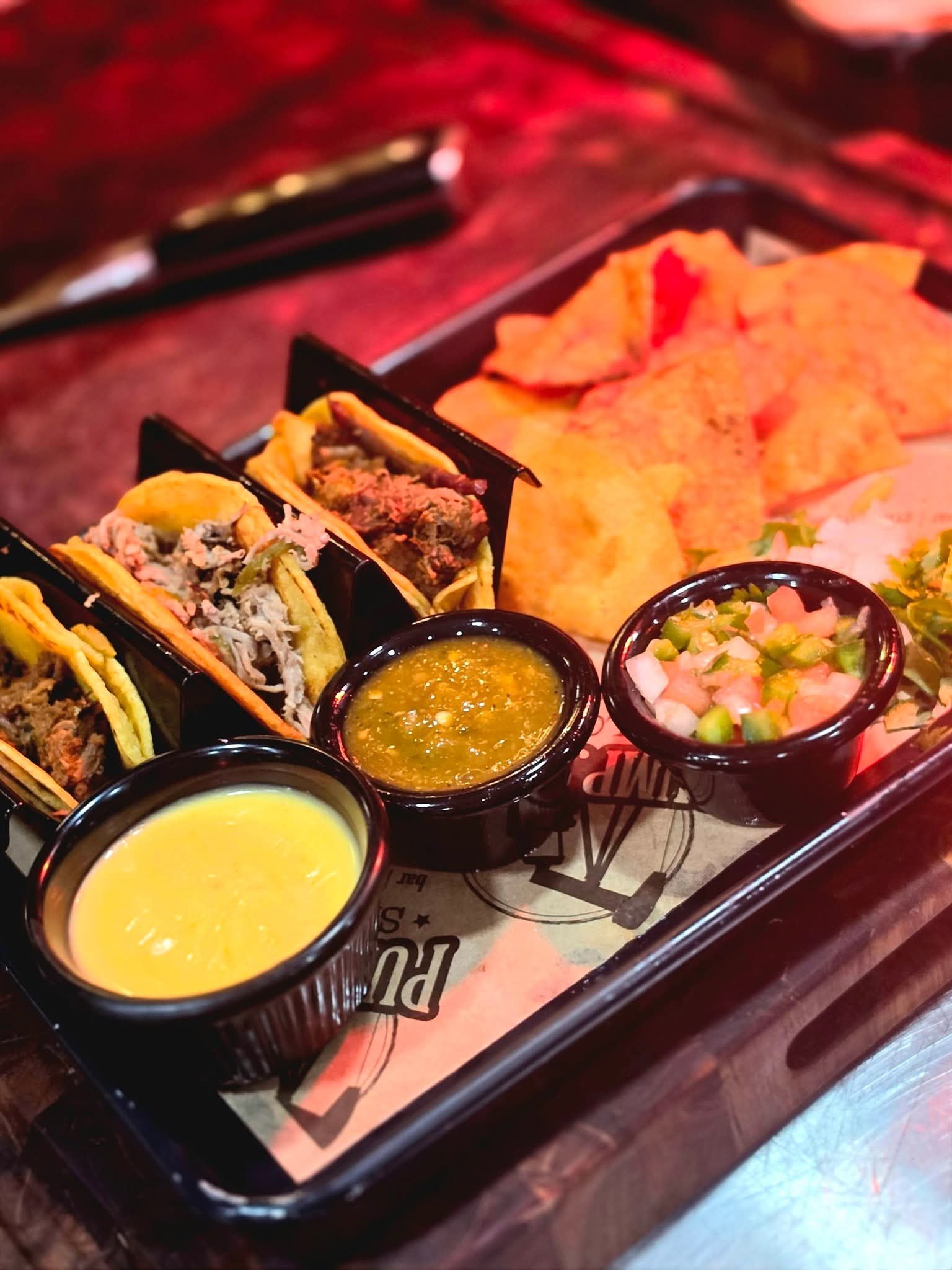 A tray of Mexican food including tacos, nachos, salsa, and dip, served on a black tray with cups of yellow sauce and salsa.