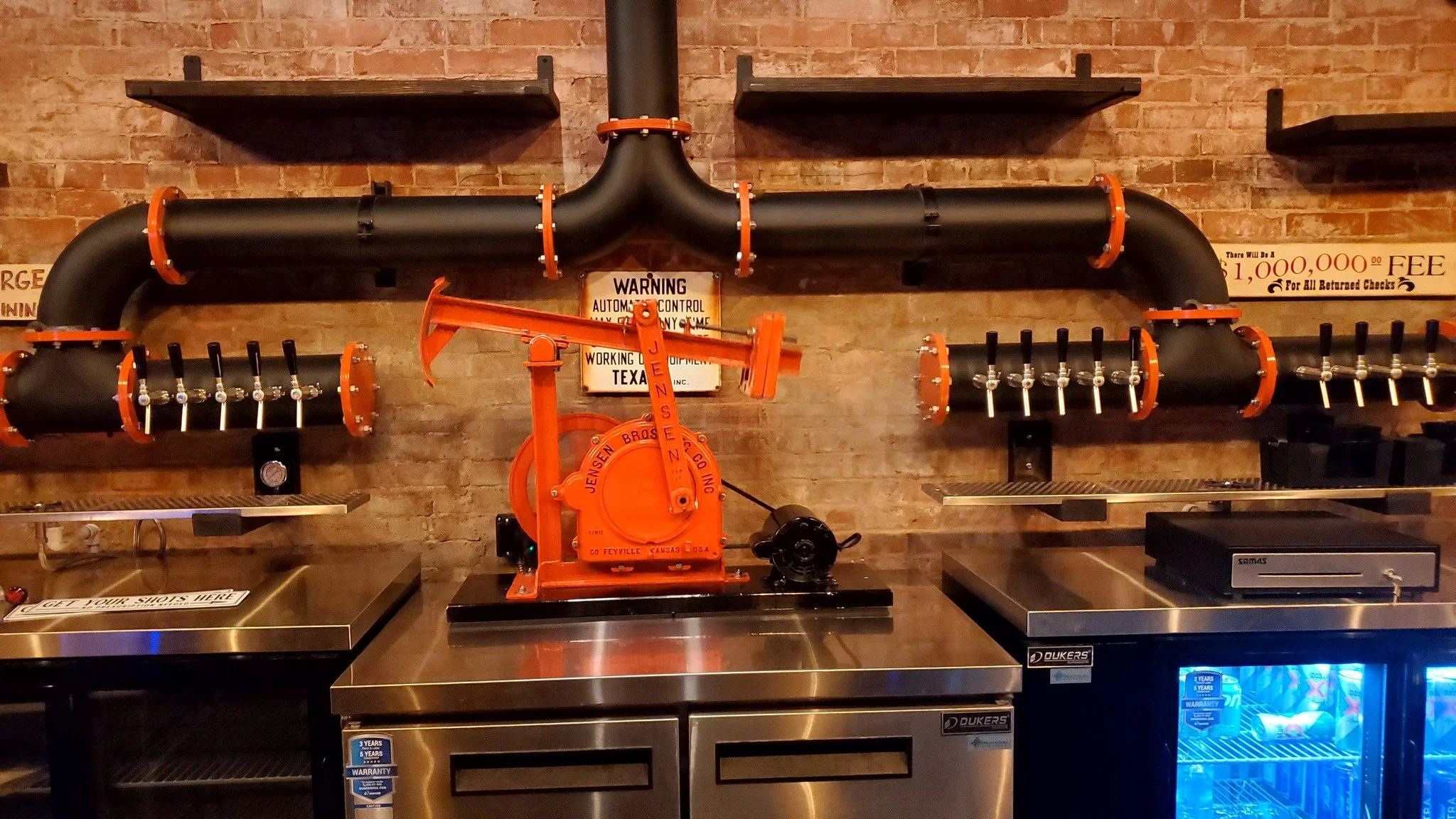 Large black industrial pipe with orange flanges and valves, mounted on a brick wall. A vintage orange hand pump labeled 'Jensen Bros Co., Inc., Feyville, Kansas, USA' is positioned in front of the pipe system.