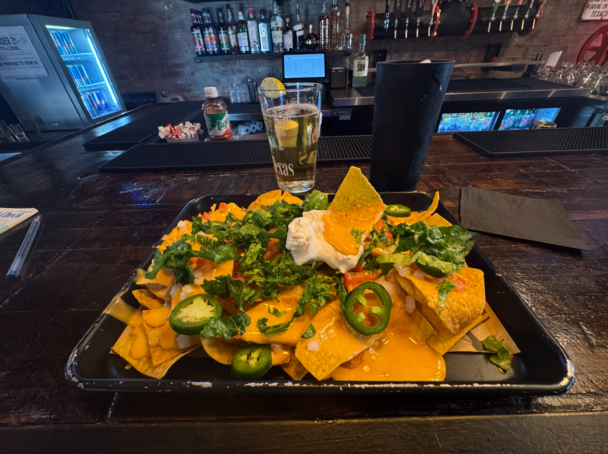 A plate of nachos topped with melted cheese, jalapeños, lettuce, sour cream, and guacamole, served on a black tray at a bar counter with drinks and bar supplies in the background.