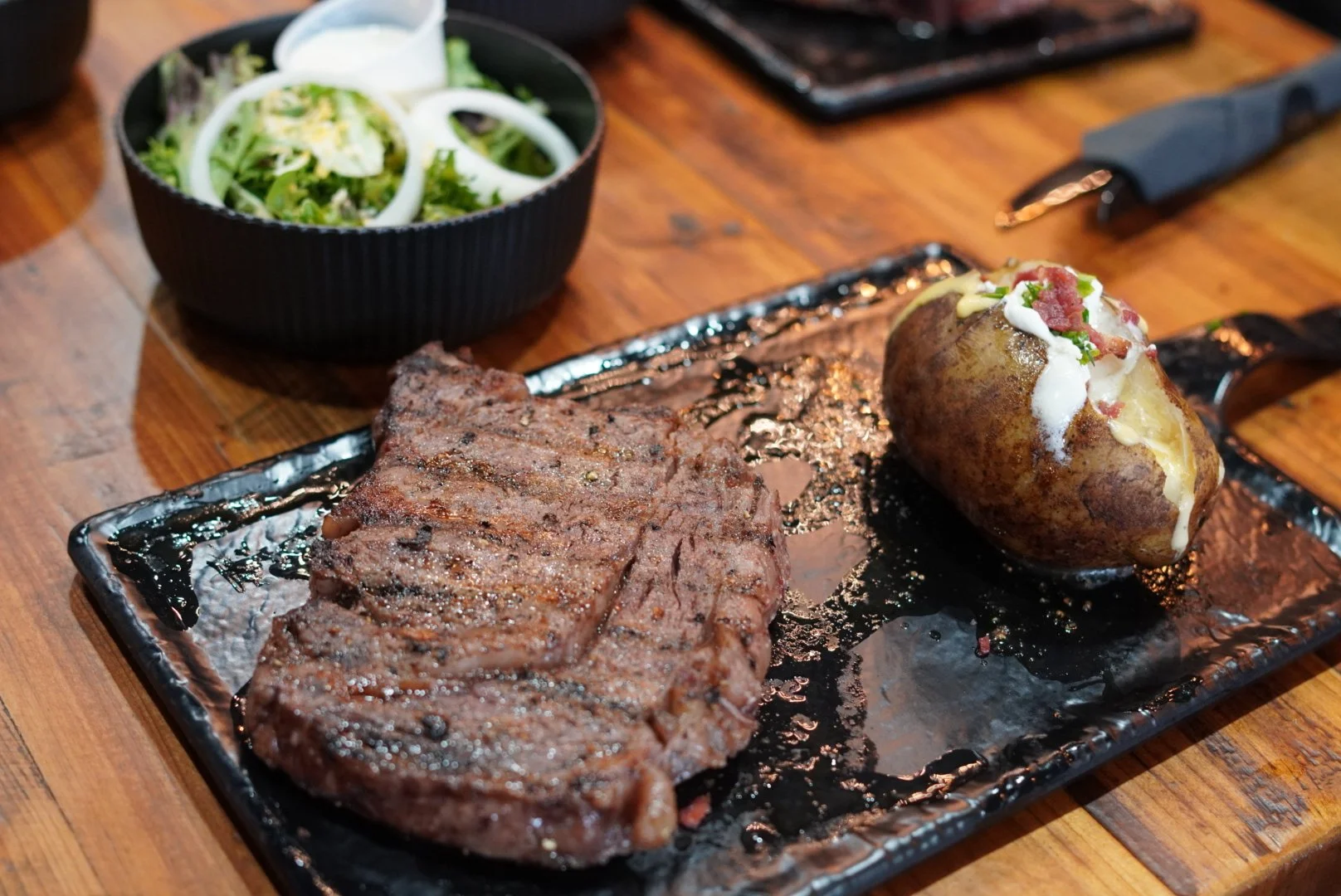 Grilled steak and baked potato with sour cream, bacon bits, and green onions on a black tray with a side salad in a black bowl