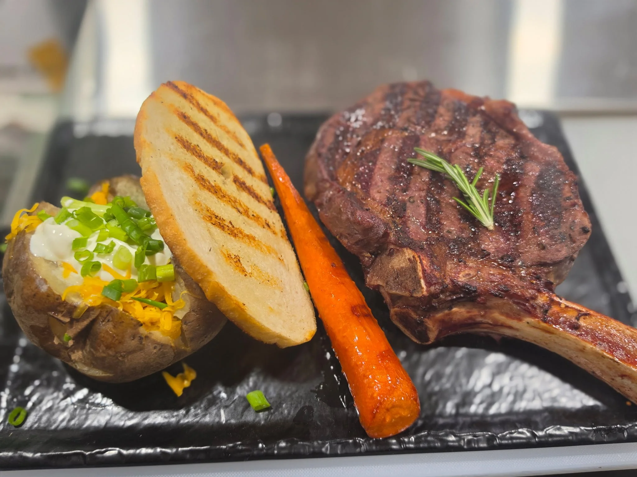 Grilled steak with a sprig of rosemary, grilled potato topped with chopped green onions, grilled carrots, and a baked potato with sour cream and green onions on a black plate.