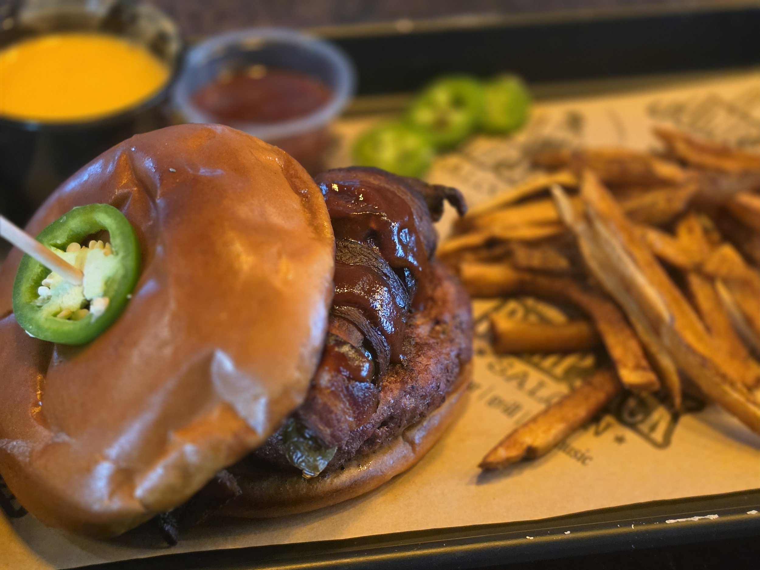 A loaded cheeseburger with lettuce, tomato, and sliced jalapeños, served with fries, and two small containers of dipping sauces in the background.