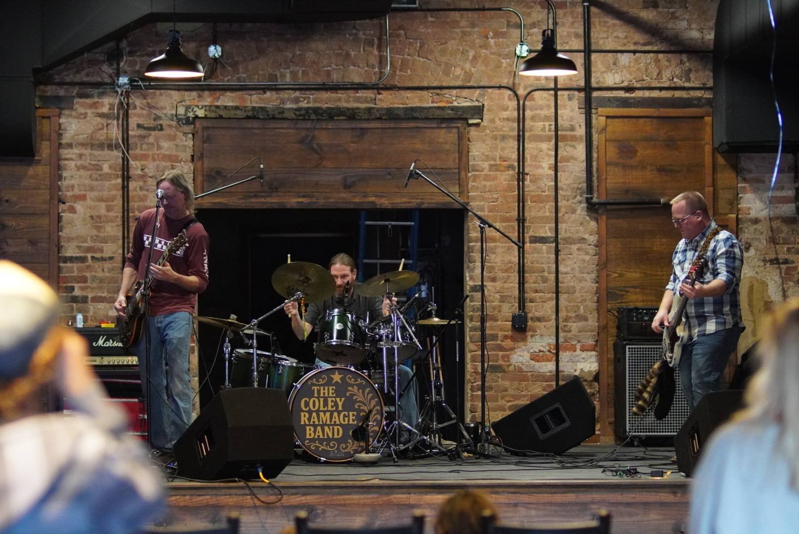 A band performing on stage with a brick wall background, consisting of a guitarist, drummer, and bassist. The drum kit has a sign that reads 'The COLEY RAMAGE BAND'.