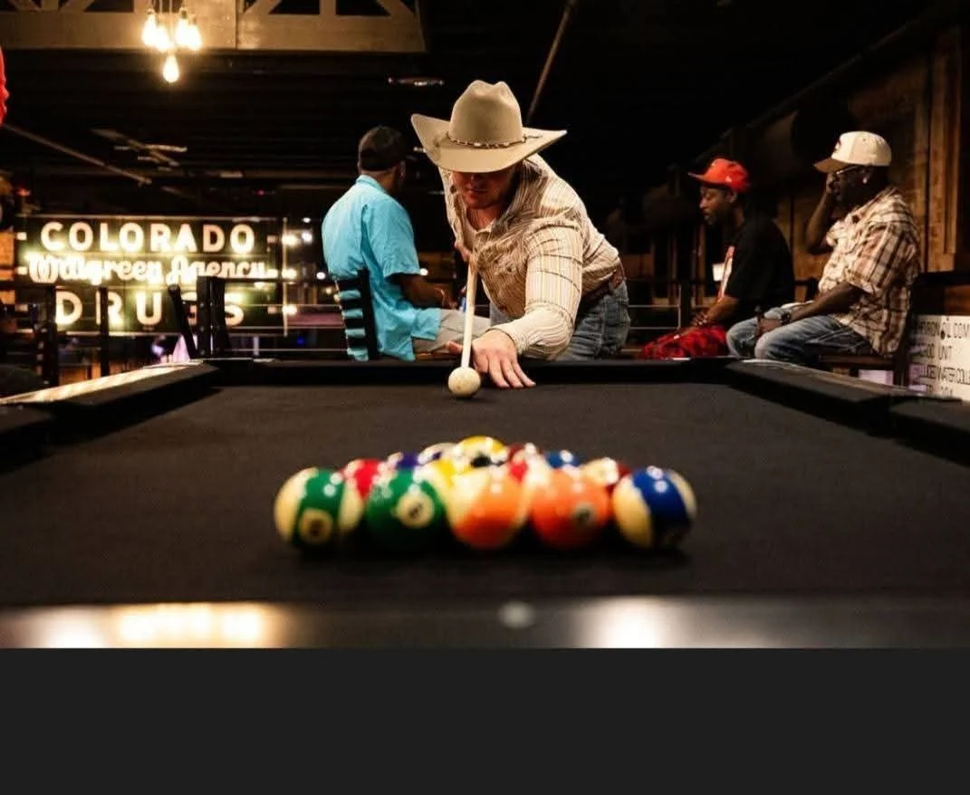 A woman in a cowboy hat aiming a cue stick at pool balls on a black pool table in a bar with other patrons.