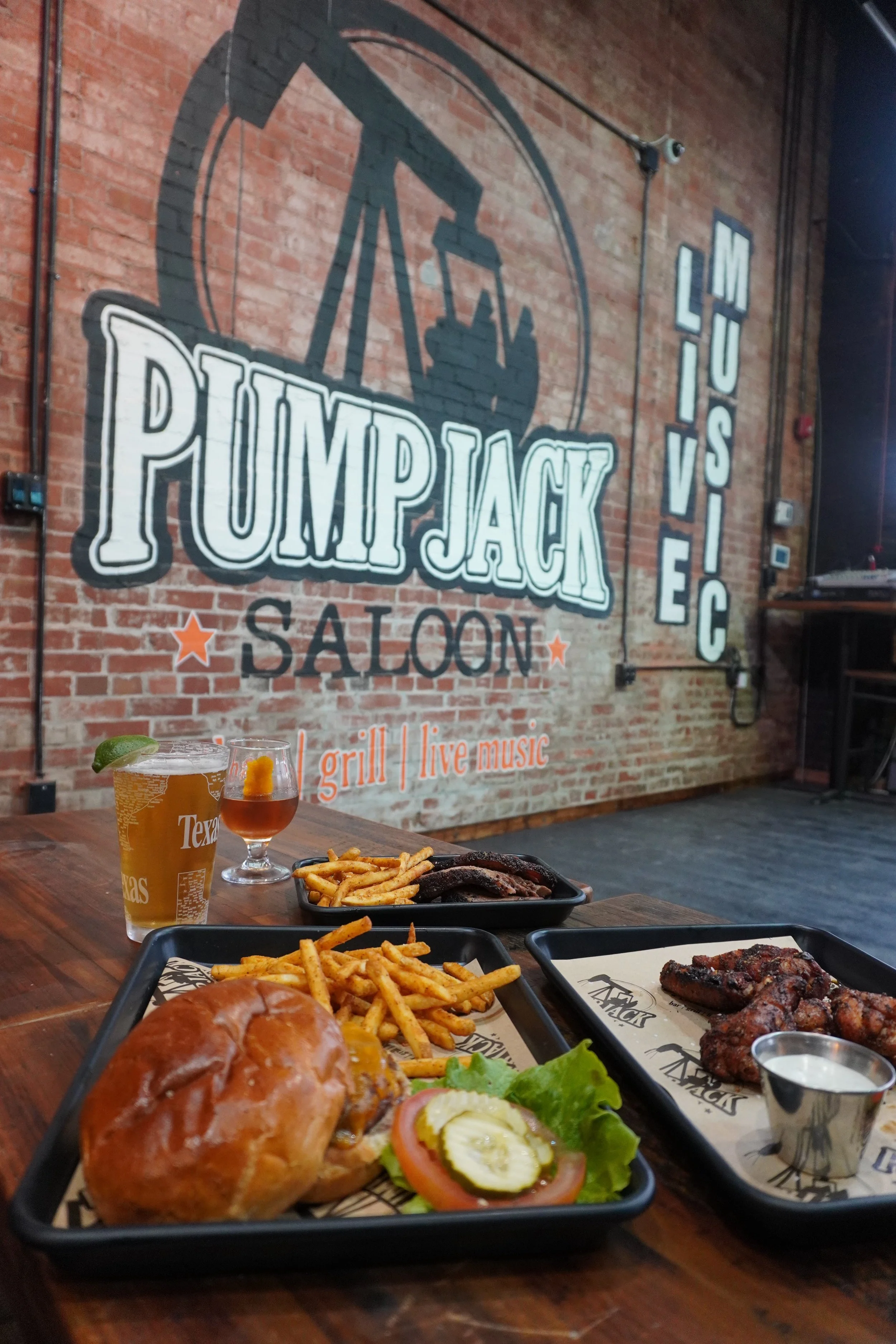 Inside a restaurant with a brick wall and a large sign reading "PUMPJACK SALOON" and “LIVE MUSIC.” The table is set with various foods including a sandwich with toppings, French fries, grilled meats, and drinks. One drink appears to be a beer with a lime wedge, and there is a glass of iced tea or soda.
