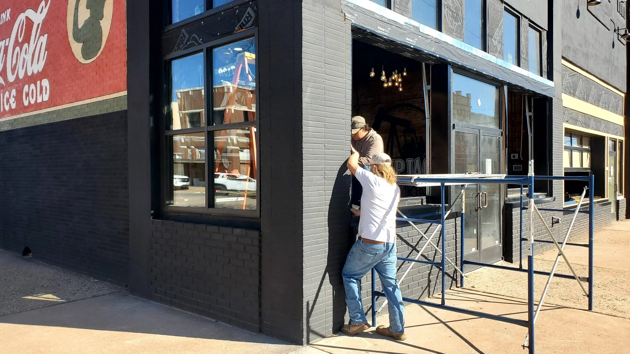 Two men working on a black brick building exterior, one standing on scaffolding and the other in a cap, installing or repairing the window.
