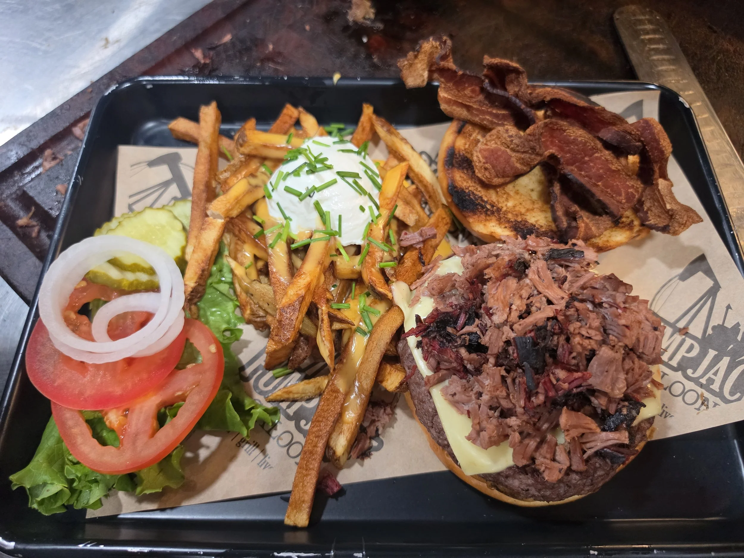 A plate with a cheeseburger topped with pulled pork and bacon, French fries with chives, sliced tomatoes, onions, pickles, and a side of shredded lettuce.