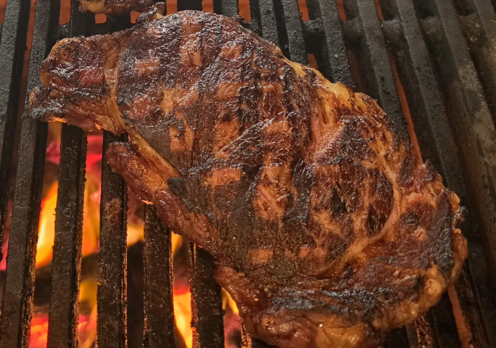 A large piece of seasoned meat, likely a steak or brisket, cooking on a hot grill over open flames.