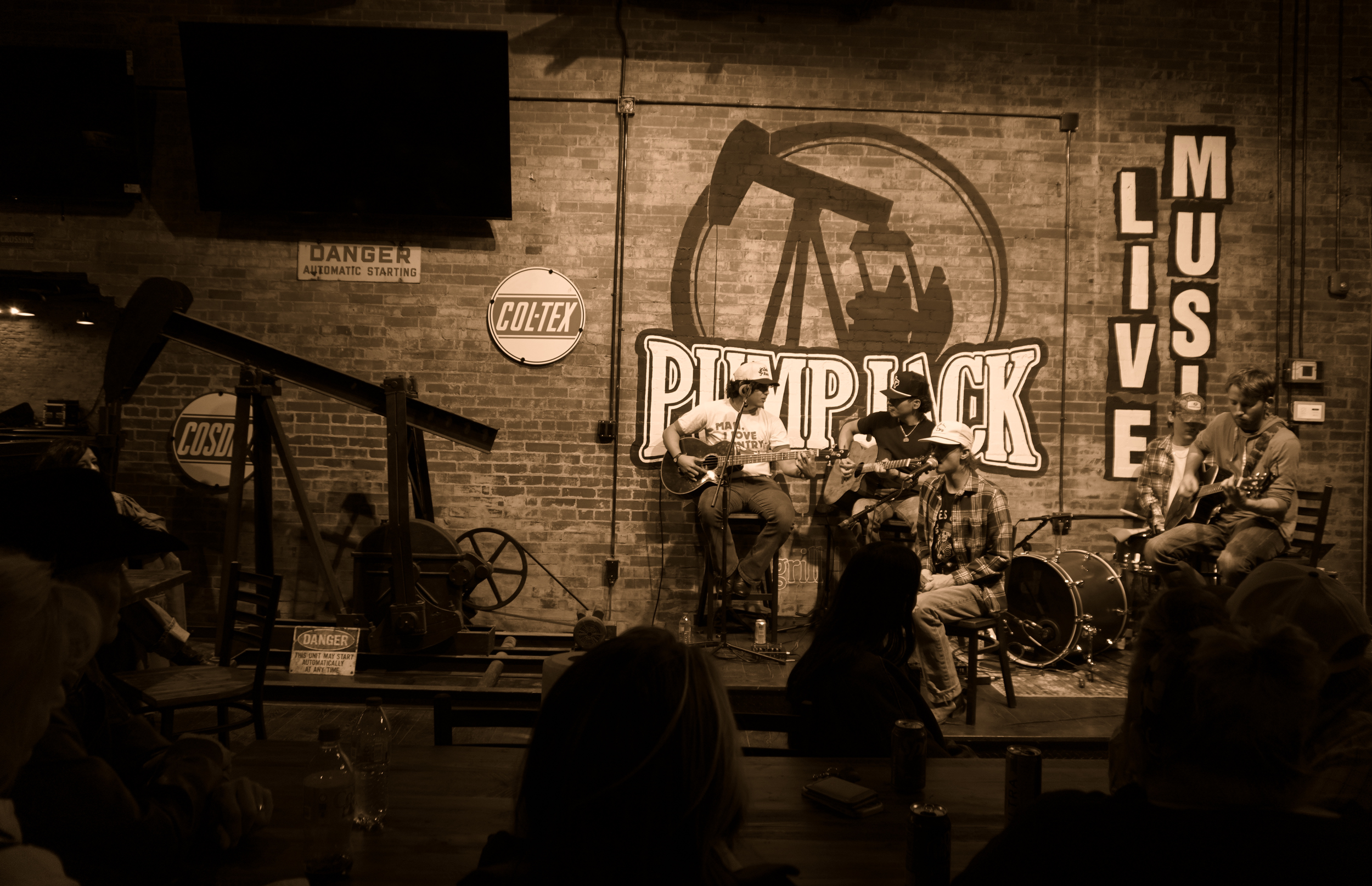 Band performing on stage at Pumpjack music venue, playing guitars and drums, audience seated at tables in dimly lit brick-walled setting.