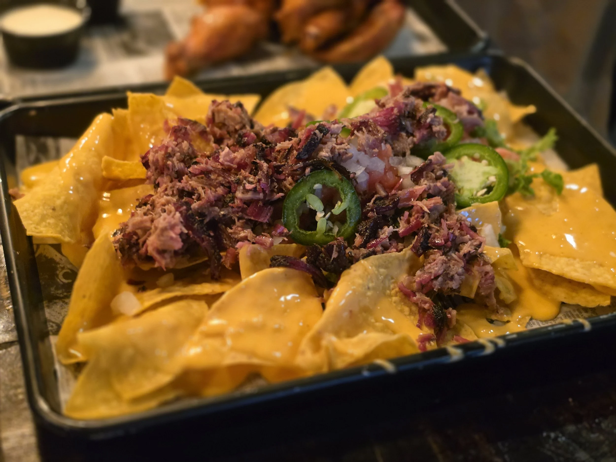 Close-up of a tray with nachos topped with shredded beef, jalapeños, and cheese, with a side of chicken wings and a small cup of what appears to be a dipping sauce or sour cream in the background.