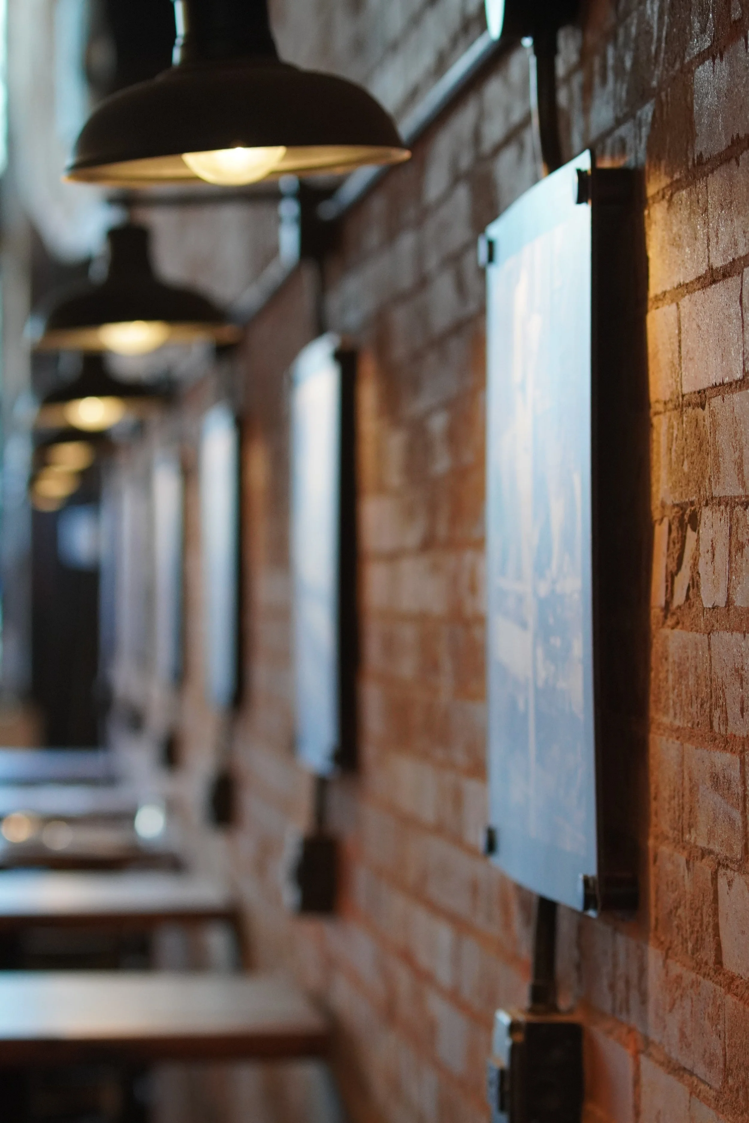 Interior view of a cafe or restaurant with exposed brick wall, black pendant lights, and blurred framed prints or menus on the wall.