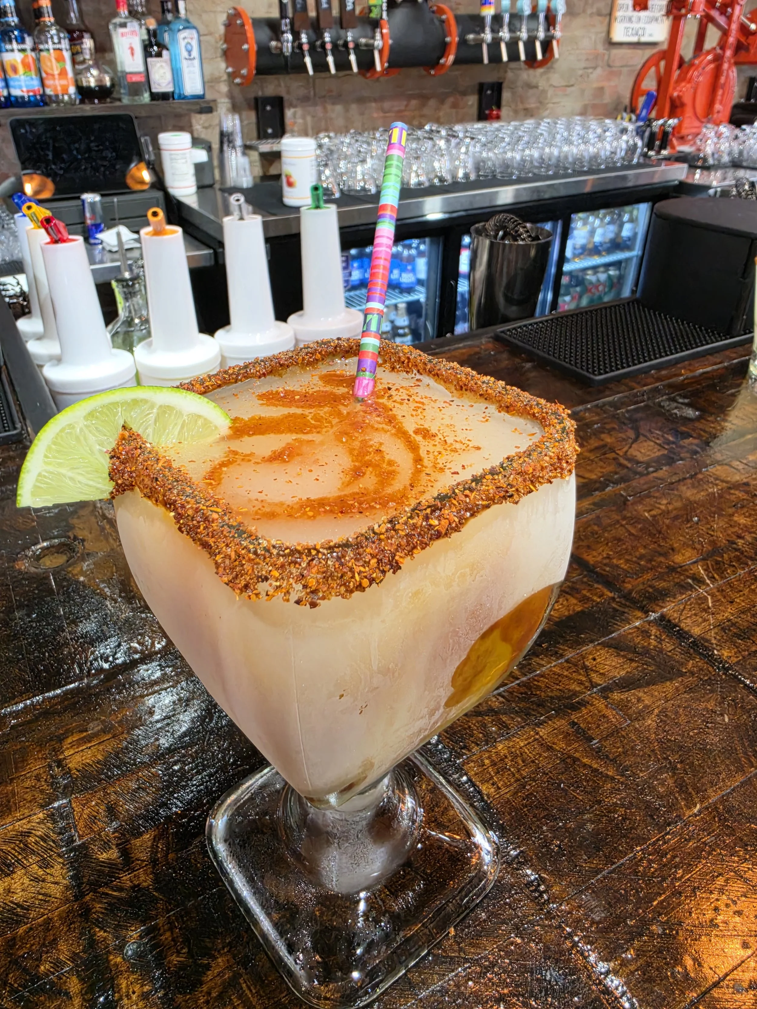 A large margarita cocktail in a glass with salt rim, lime wedge, colorful straw, on a wooden bar counter, with bar supplies and bottles in the background.
